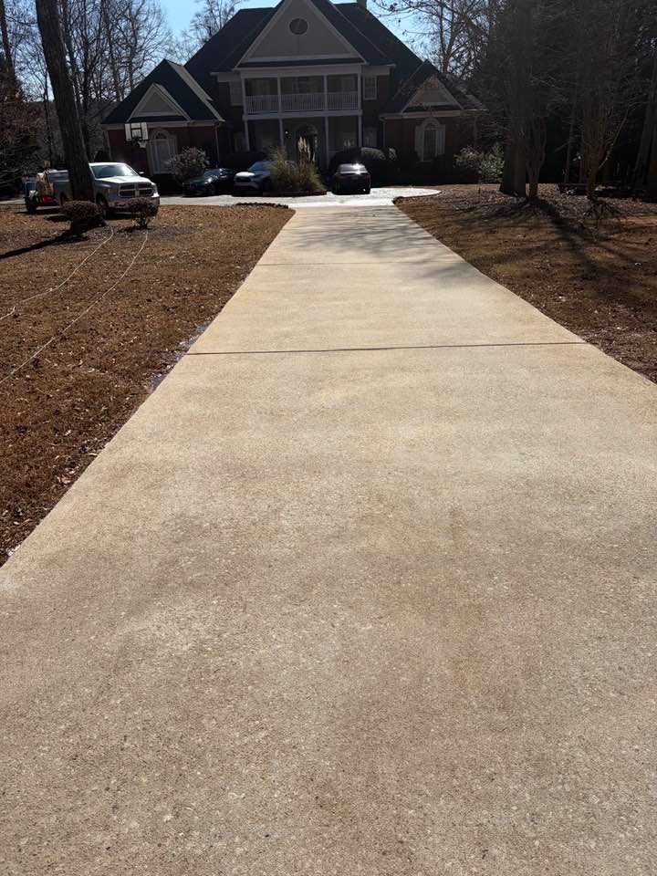 A concrete driveway leads to a large, two-story house surrounded by trees on a sunny day.
