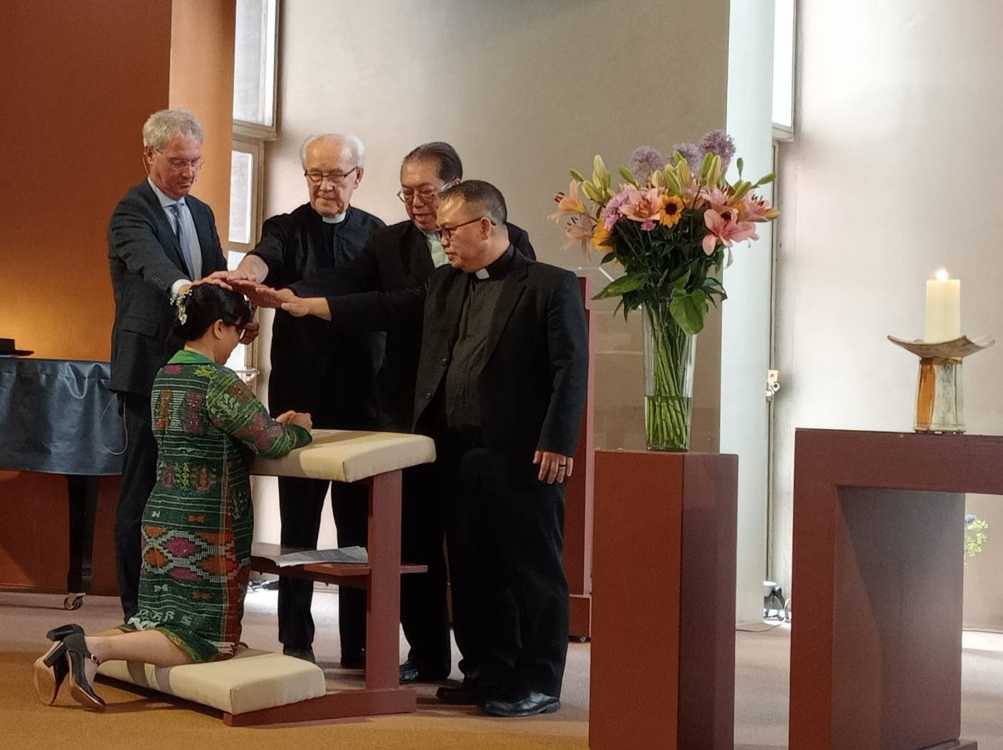 Een vrouw knielt terwijl meerdere mensen hun handen op haar hoofd leggen in een kerk; bloemen en een kaars zijn zichtbaar.