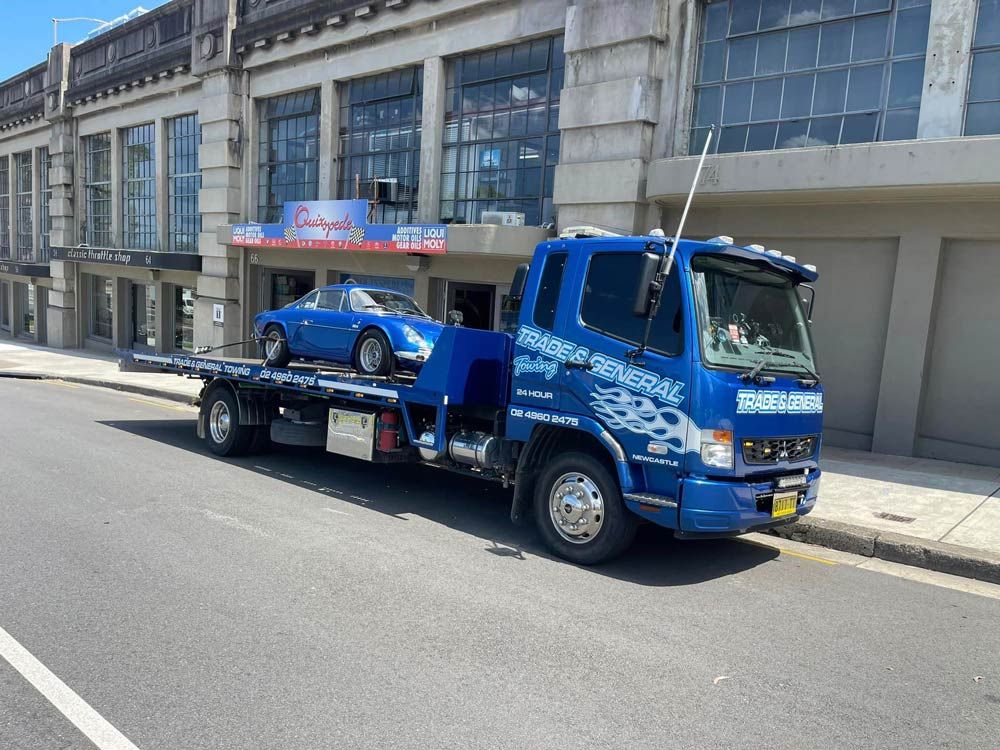 A Blue Tow Truck With a Blue Car on the Back is Parked on the Side of the Road — Trade & General Towing in Cessnock, NSW