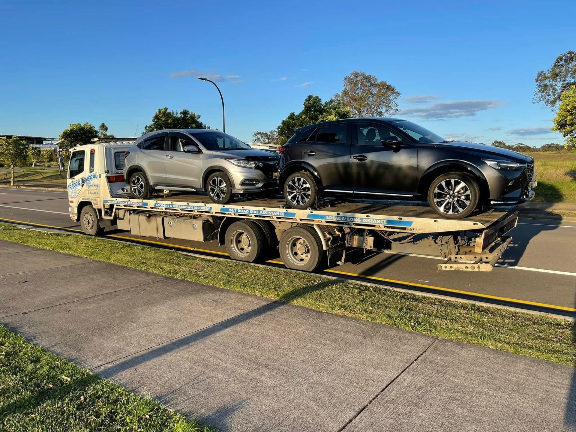 Two SUVs loaded on a car trailer being towed along a roadside in daylight