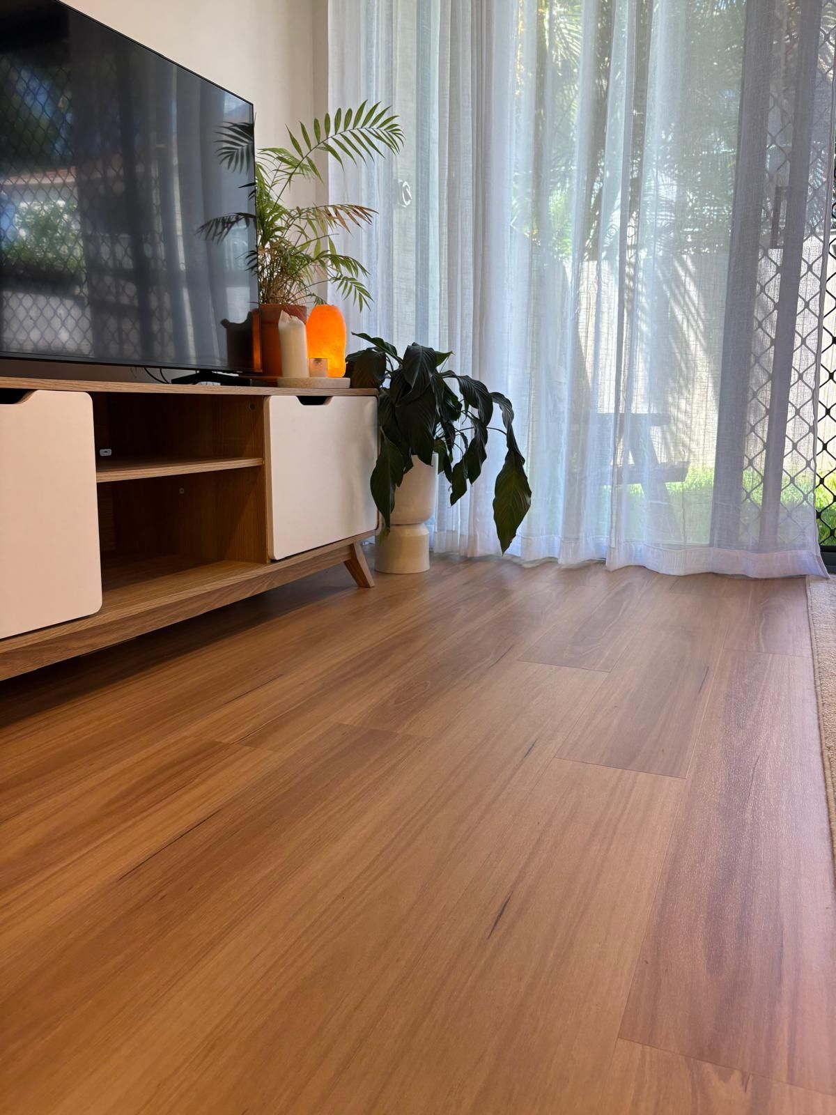 Living room with wooden floor, TV stand, potted plants, and sheer curtains by a bright window