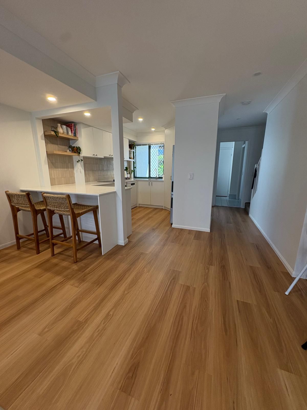 Bright empty apartment interior with wood floors, bar stools, and a kitchen opening to a hallway.