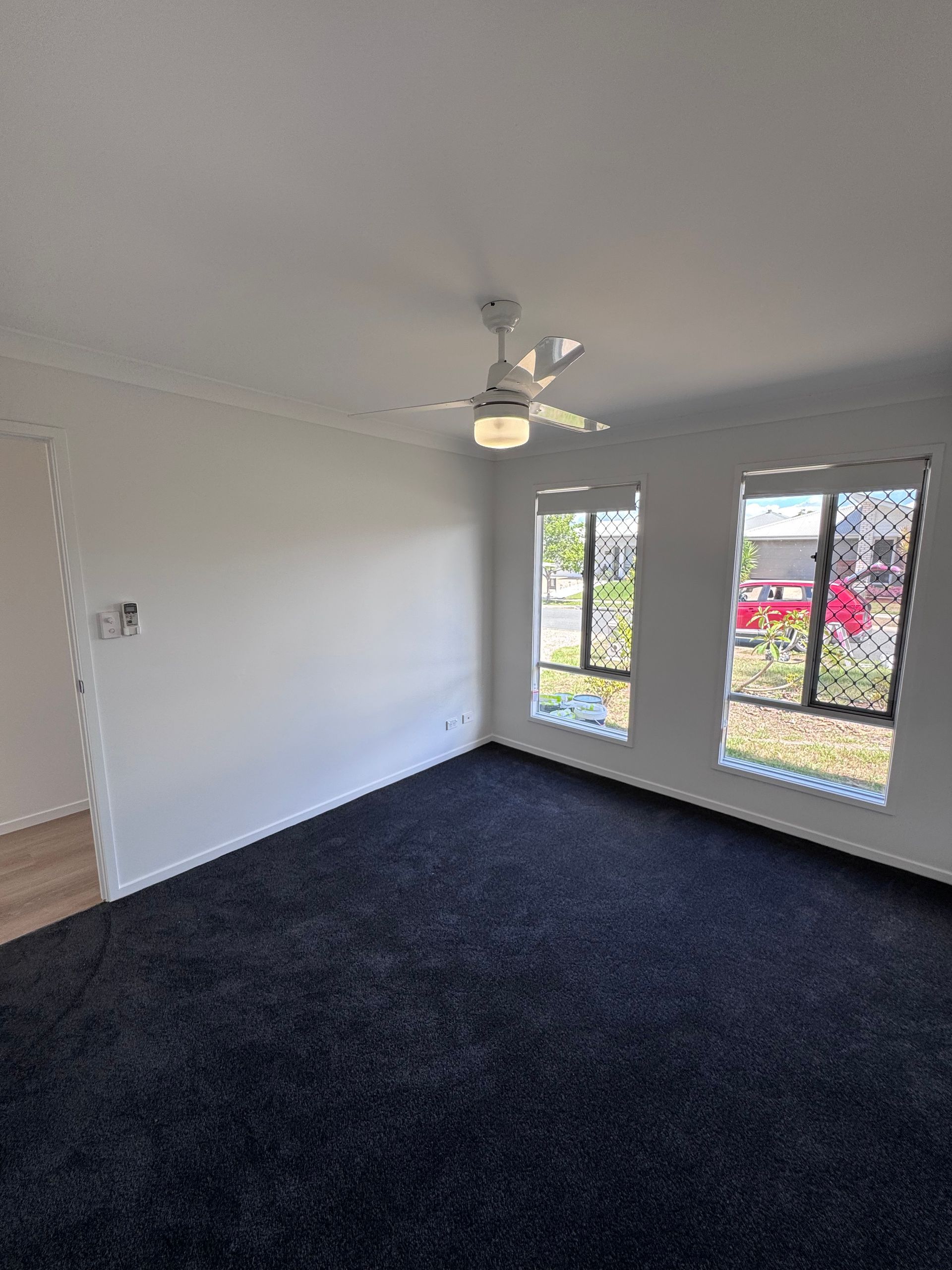 Empty living room with dark carpet, white walls, ceiling fan, and two windows overlooking greenery.