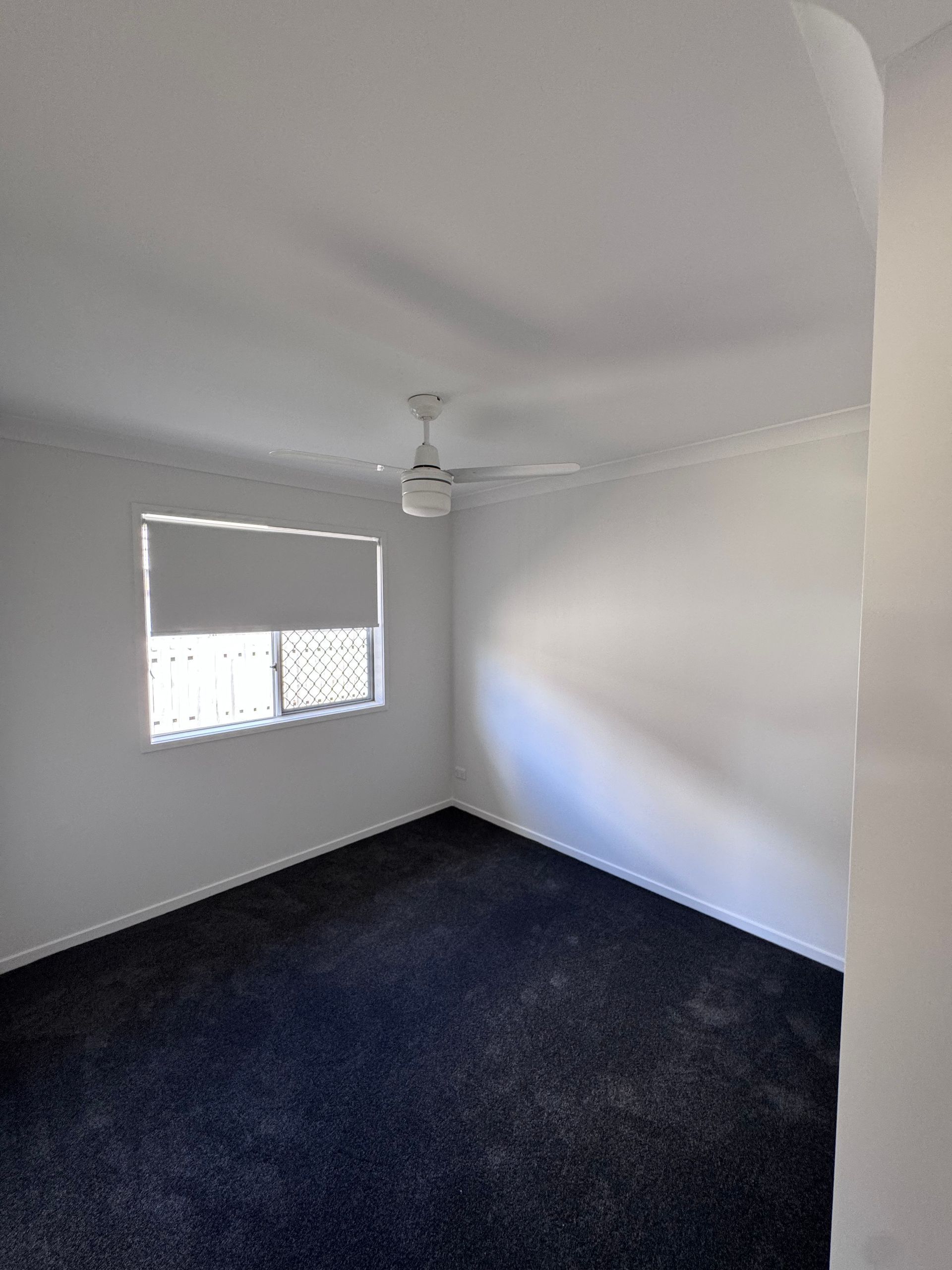 Empty white bedroom with dark carpet, a window, and a ceiling fan