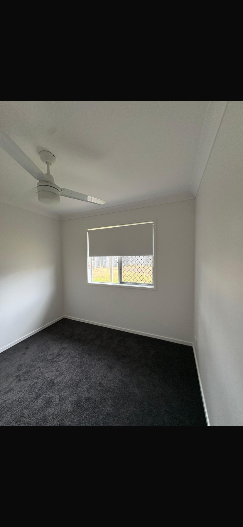 Empty bedroom with white walls, dark carpet, ceiling fan, and a window with blinds.