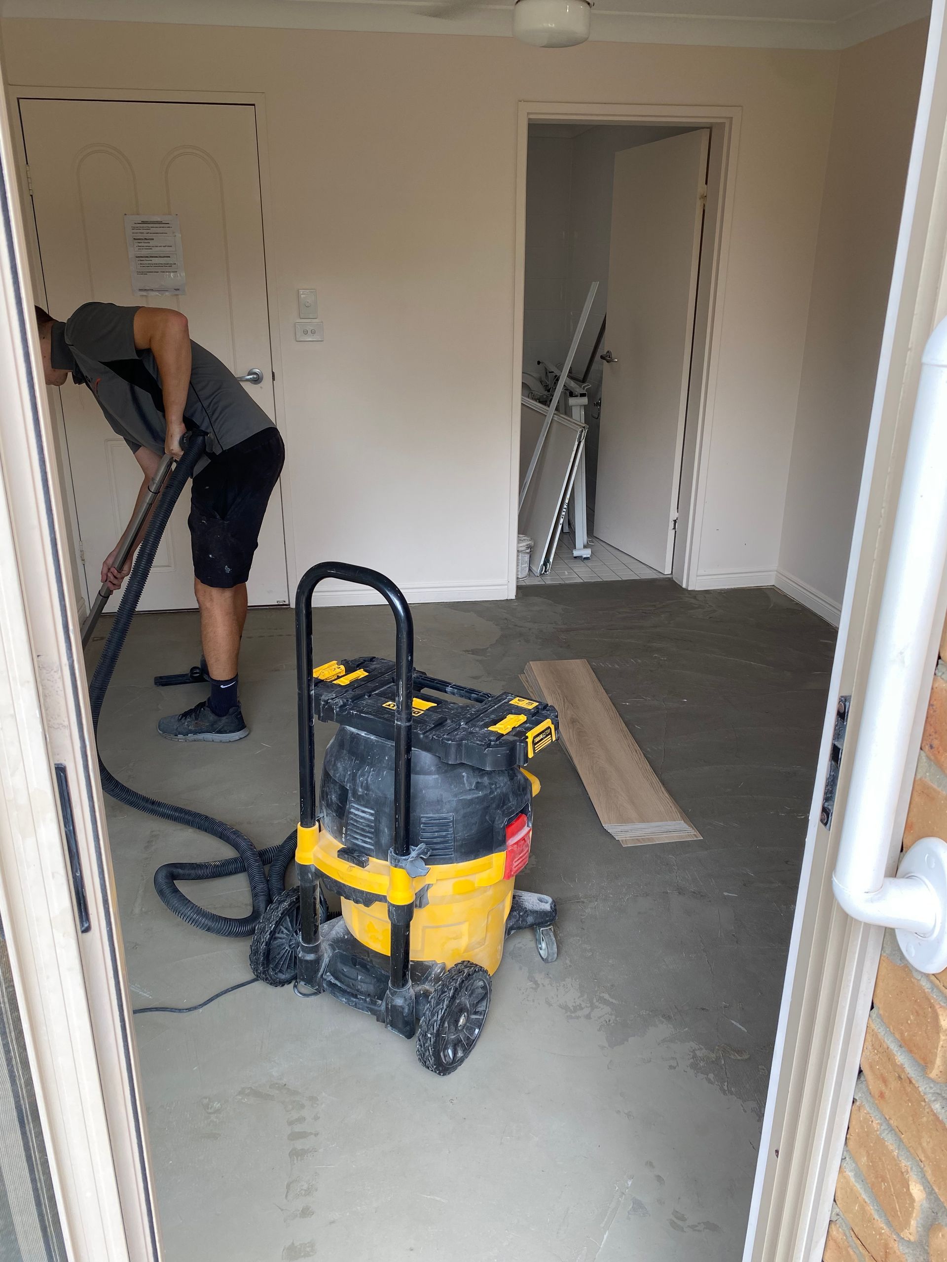 Worker vacuuming a beige room with a yellow-and-black shop vac and tools in the doorway