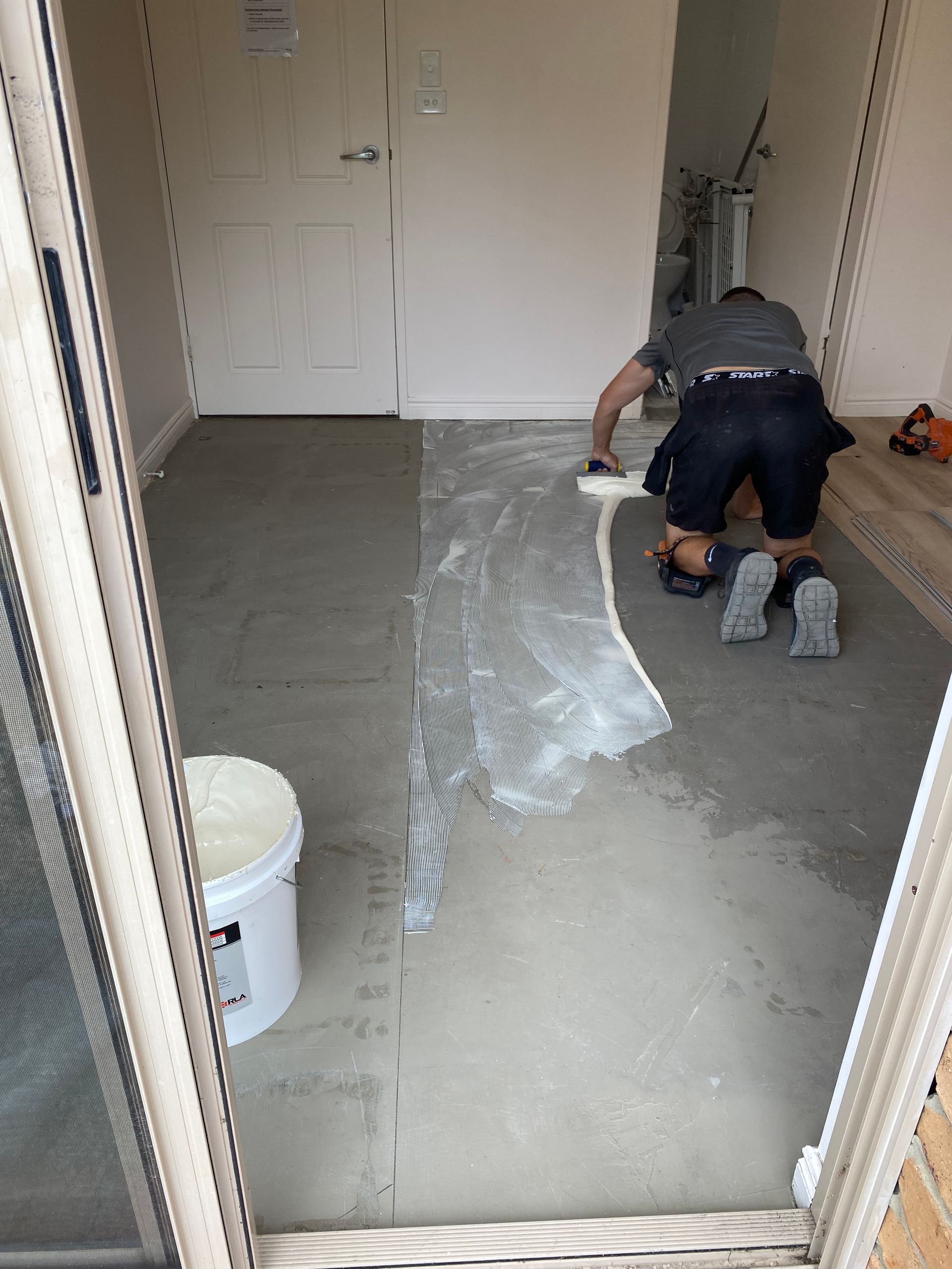 Person kneels on a concrete floor, spreading white material in a narrow hallway under construction.