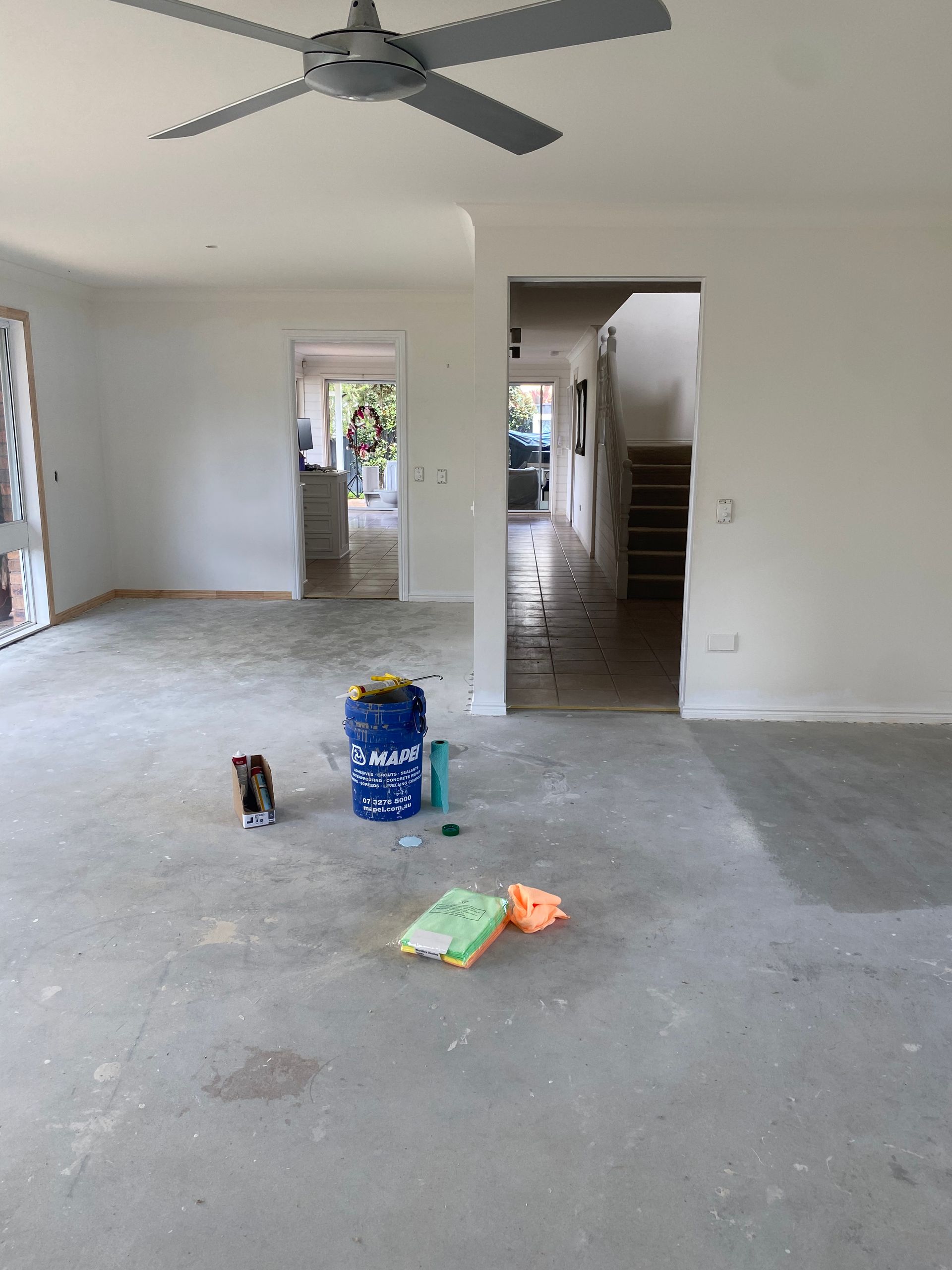 Empty living room with gray floor, ceiling fan, stairs, and a few paint supplies on the floor