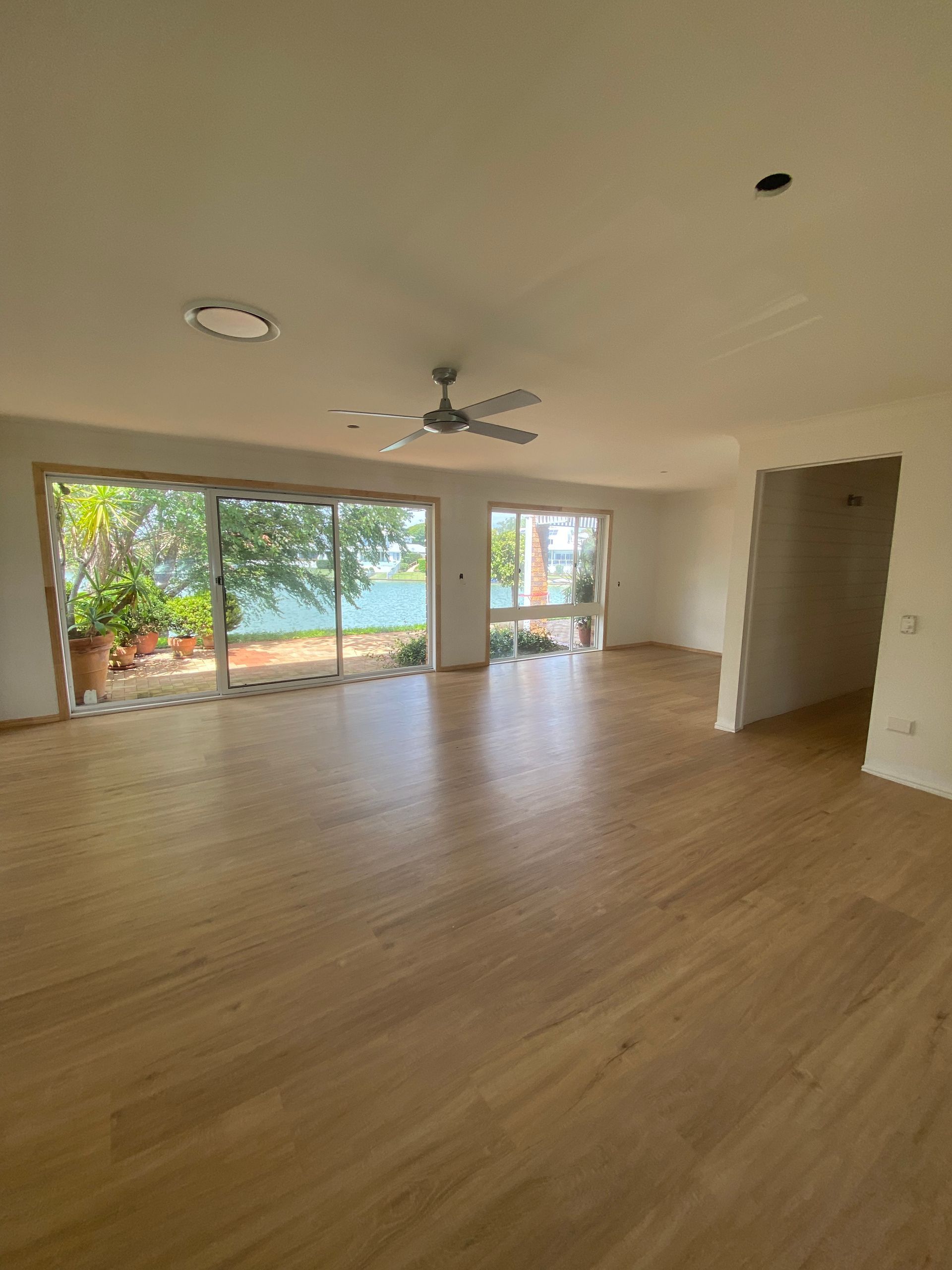 Empty living room with wood floors, ceiling fan, and large sliding glass doors opening to a leafy patio.