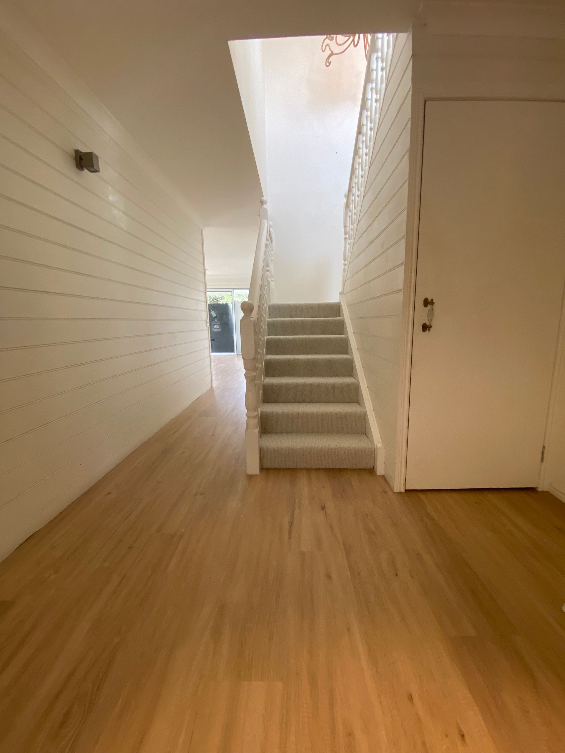 Empty hallway with light wood floor, white walls, and a staircase leading up to a bright doorway