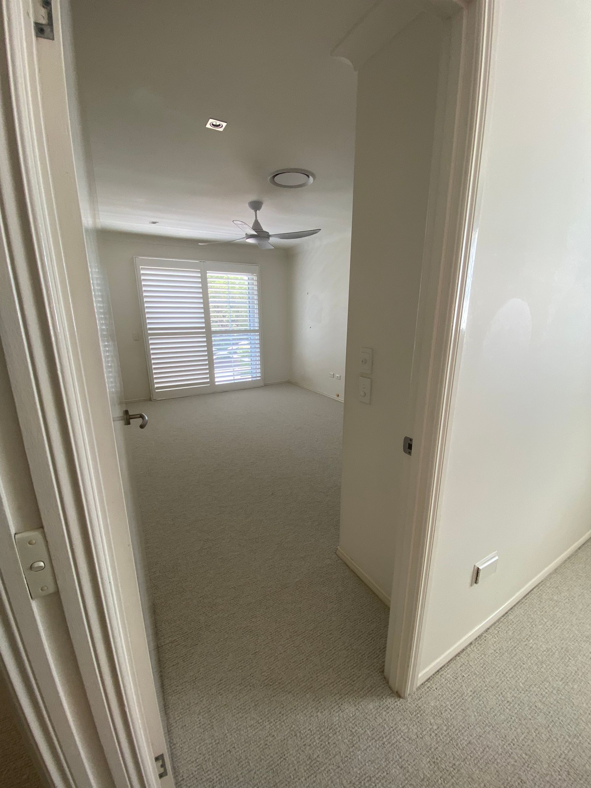 Empty carpeted hallway and bedroom with white walls, window blinds, and ceiling fans.