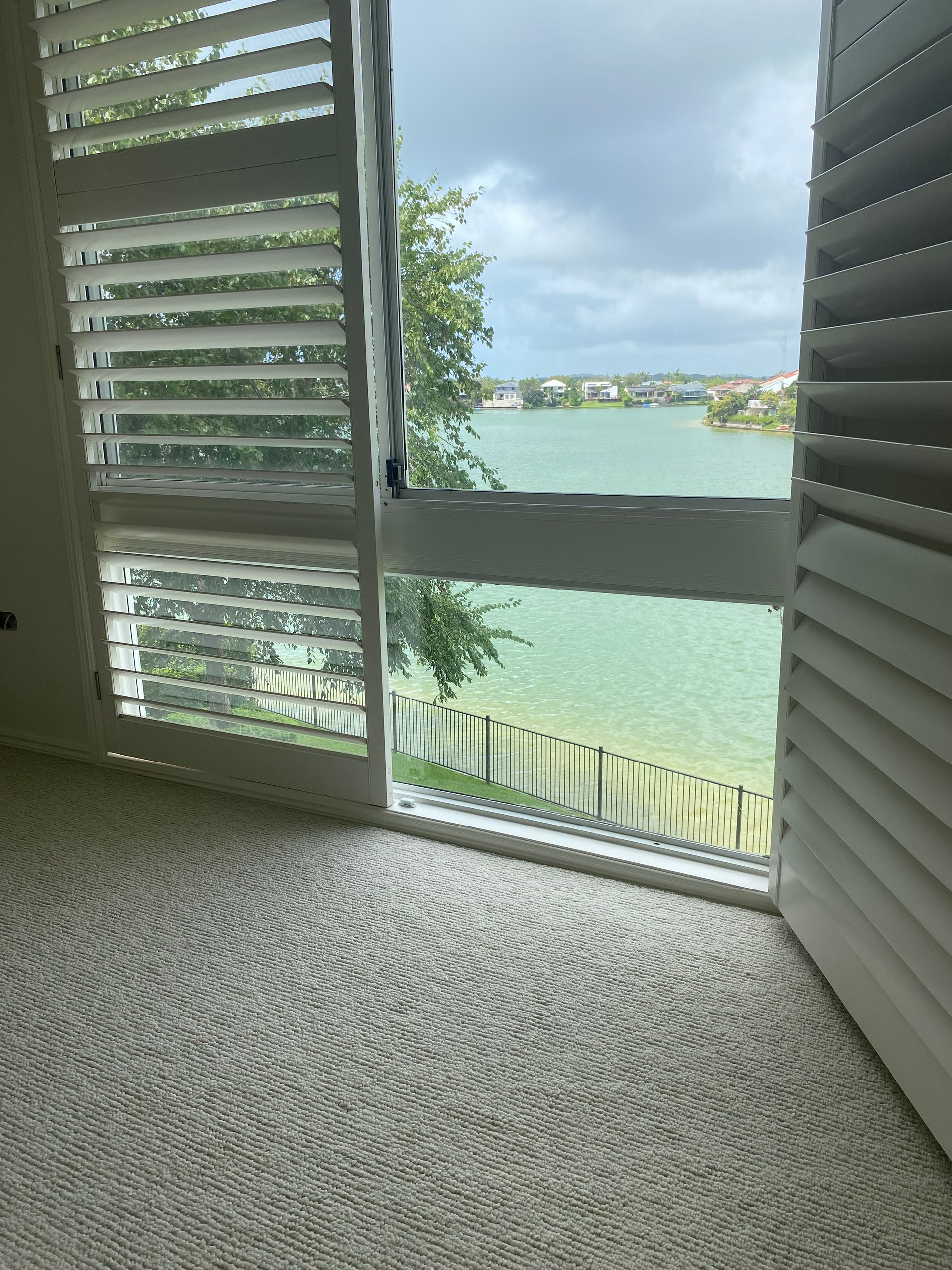 Open white shutters framing a bright waterfront view with green-blue water and a grassy shore.
