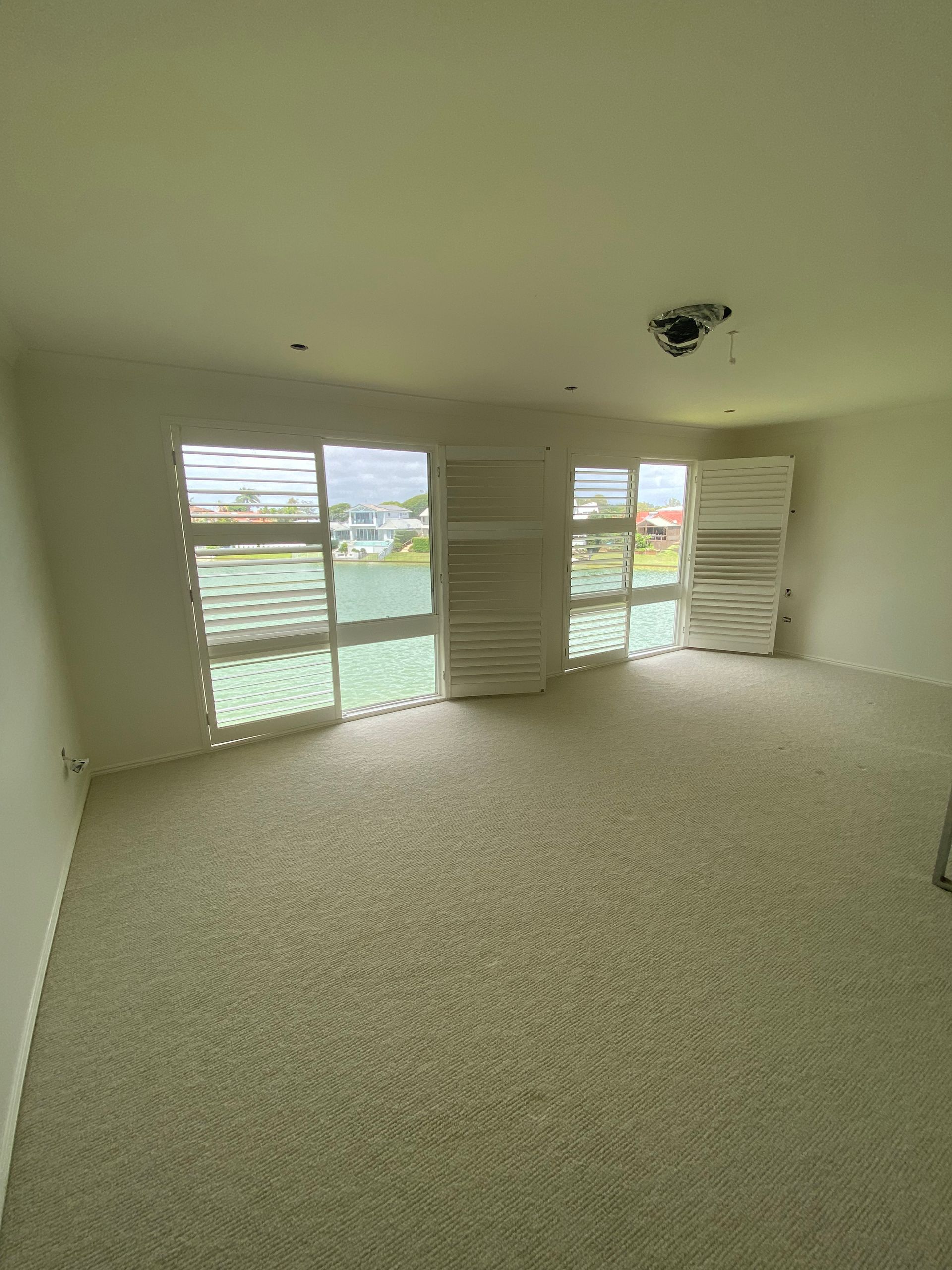Empty carpeted room with white shutters and large windows overlooking water