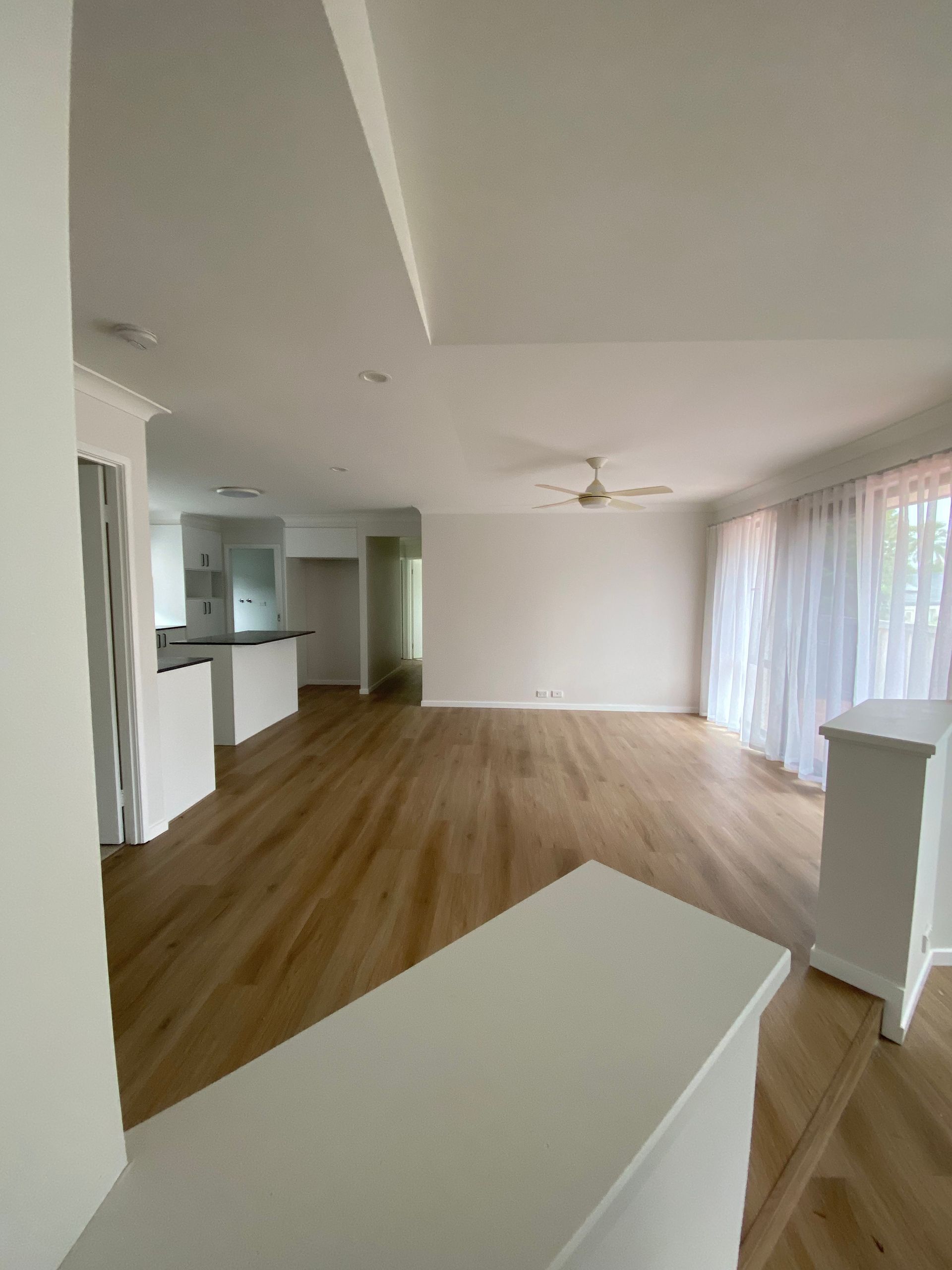Empty modern living room with wood floors, white walls, and large windows with sheer curtains.