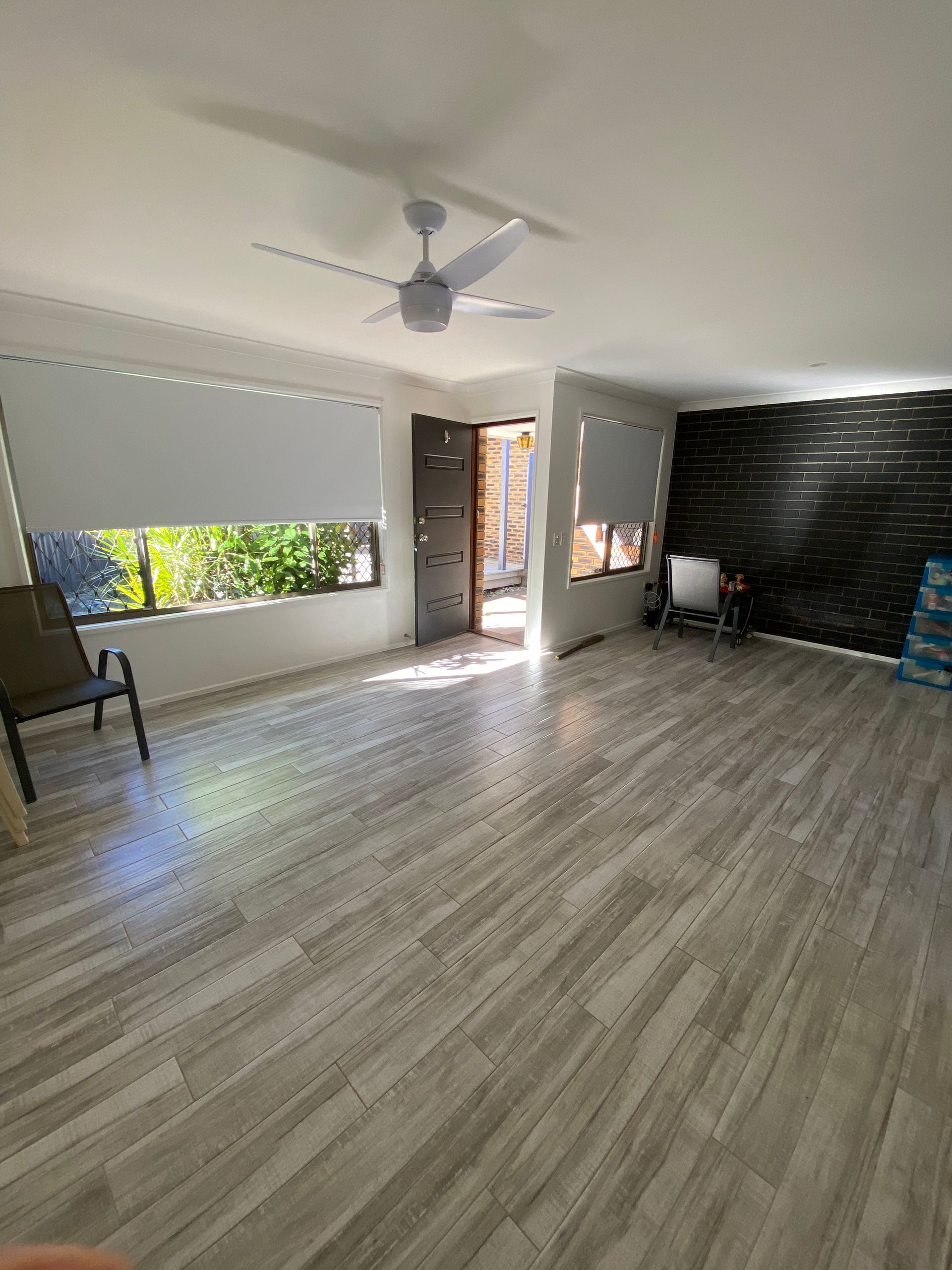 Empty modern living room with gray wood-look floors, ceiling fan, large window, and open front door.