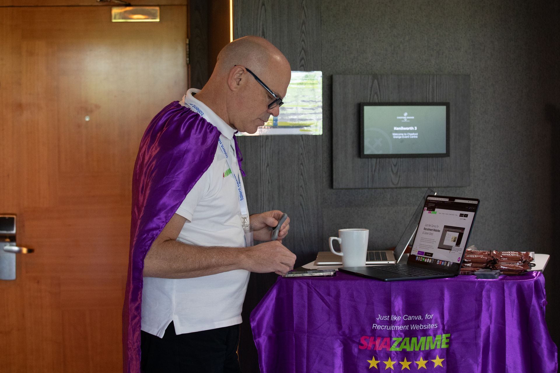 A man in a purple cape is standing in front of a table with a laptop on it.