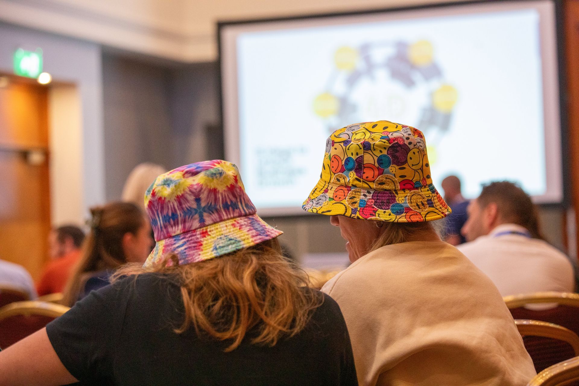Two people wearing colorful hats are sitting in front of a projector screen.