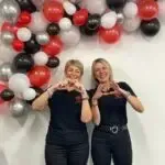 Two women are making a heart shape with their hands in front of a wall of balloons.