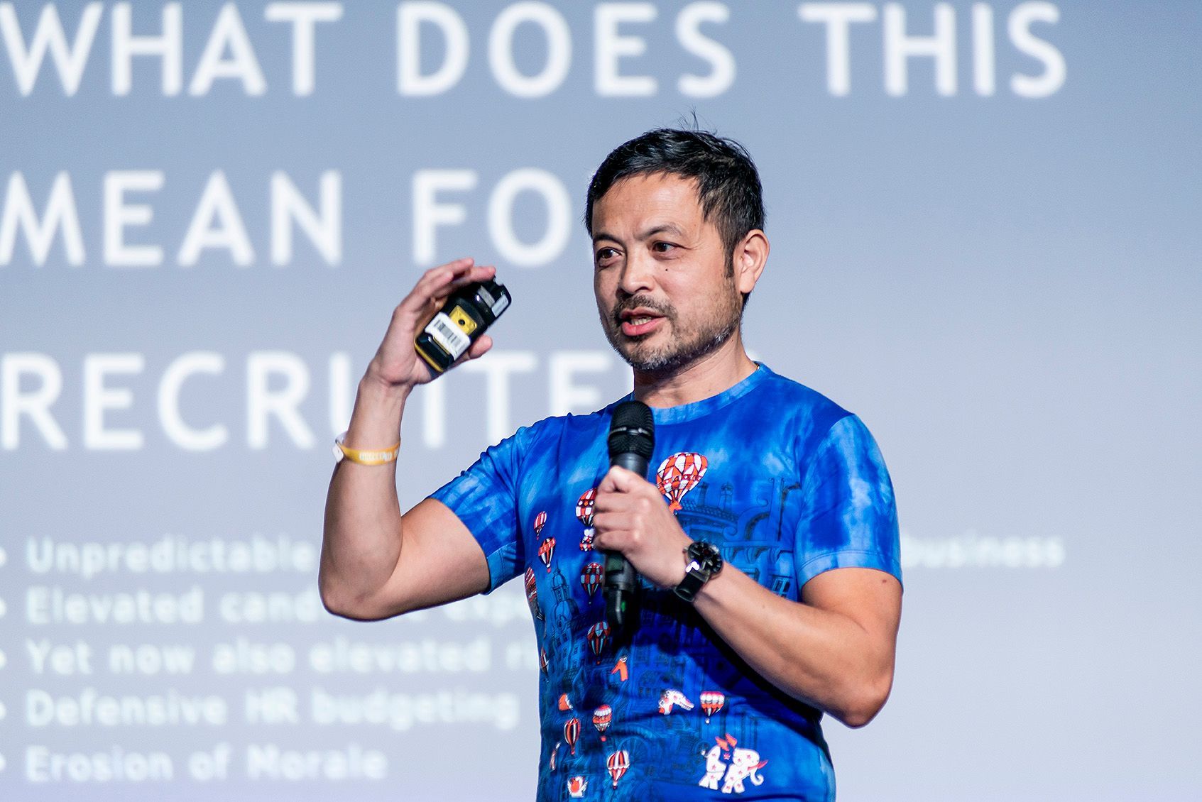 A man is holding a microphone in front of a screen that says what does this mean for recruiters