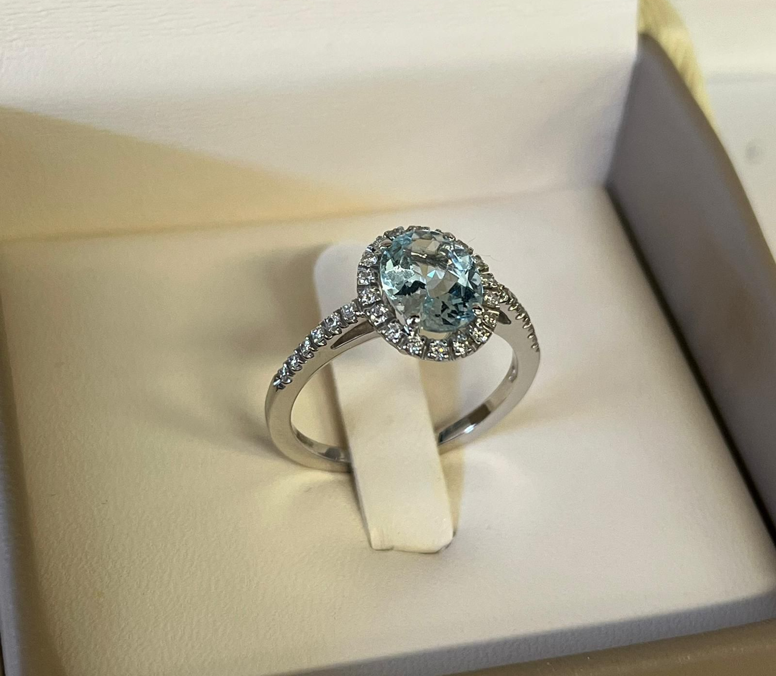 Anello in argento con pietra preziosa ovale blu e alone di diamanti, presentato in una scatola per anelli bianca.