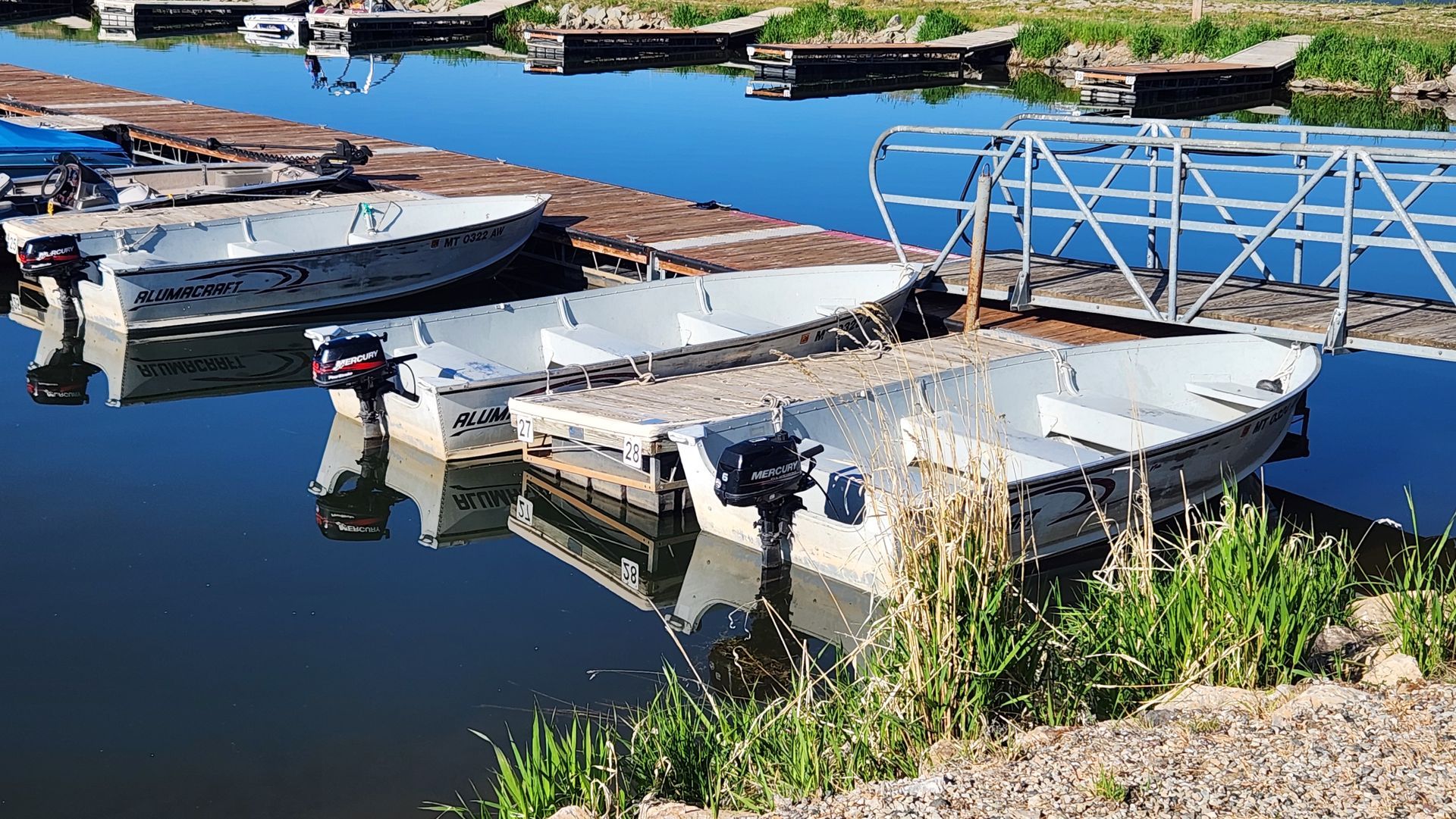 Small boats moored at a calm marina with a metal dock and blue water reflections