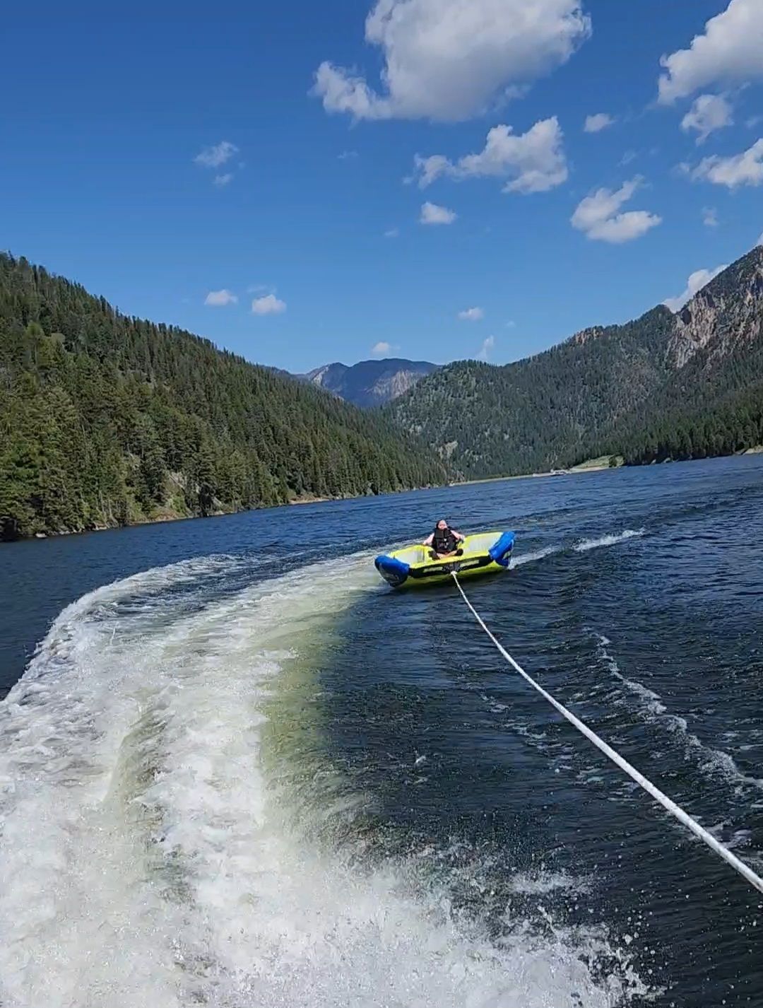 Person on yellow tube being towed across a blue lake, with forested mountains and a sunny sky.