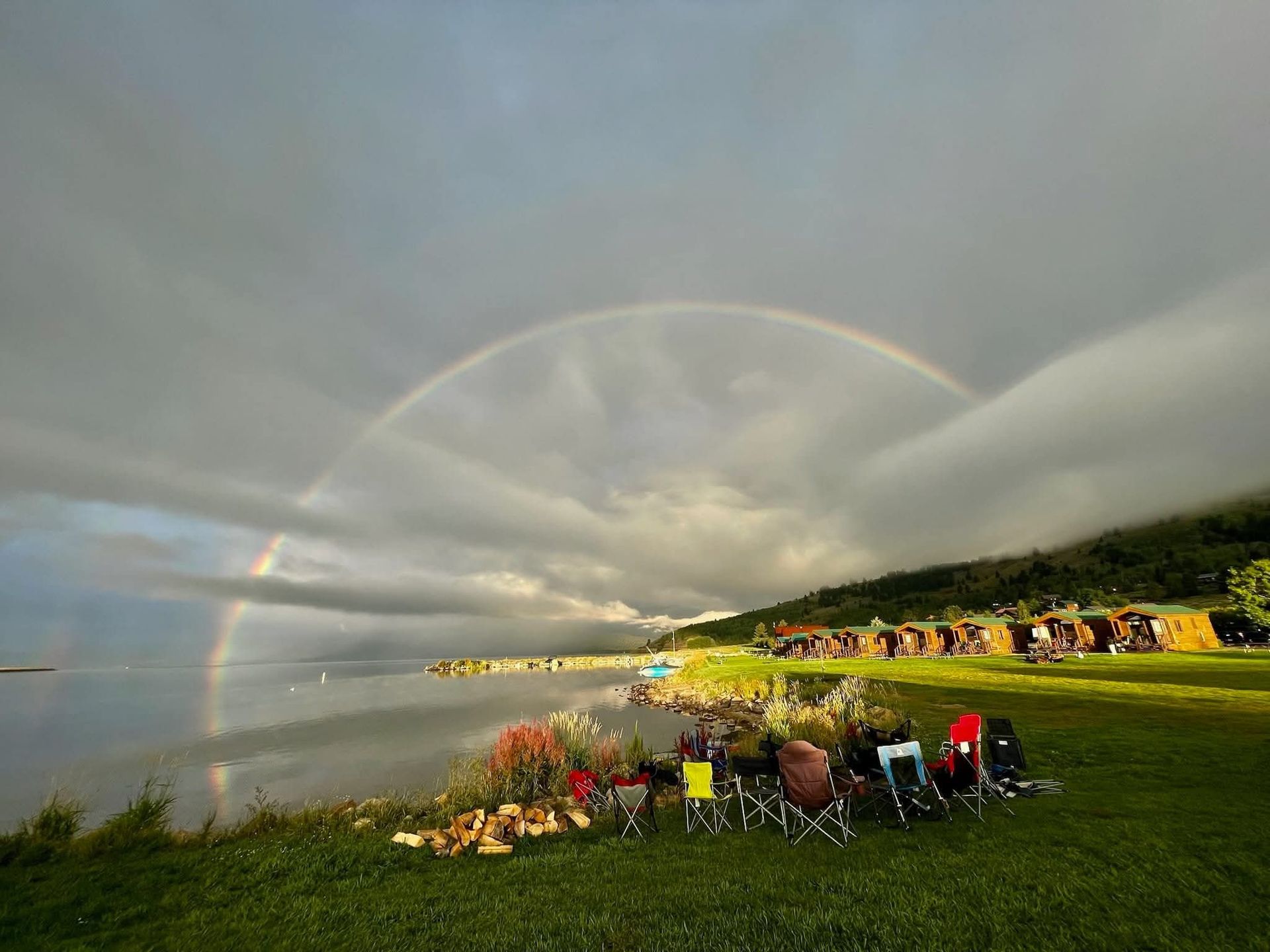 Double rainbow over a lakeside gathering with chairs, flowers, and green hills at sunset