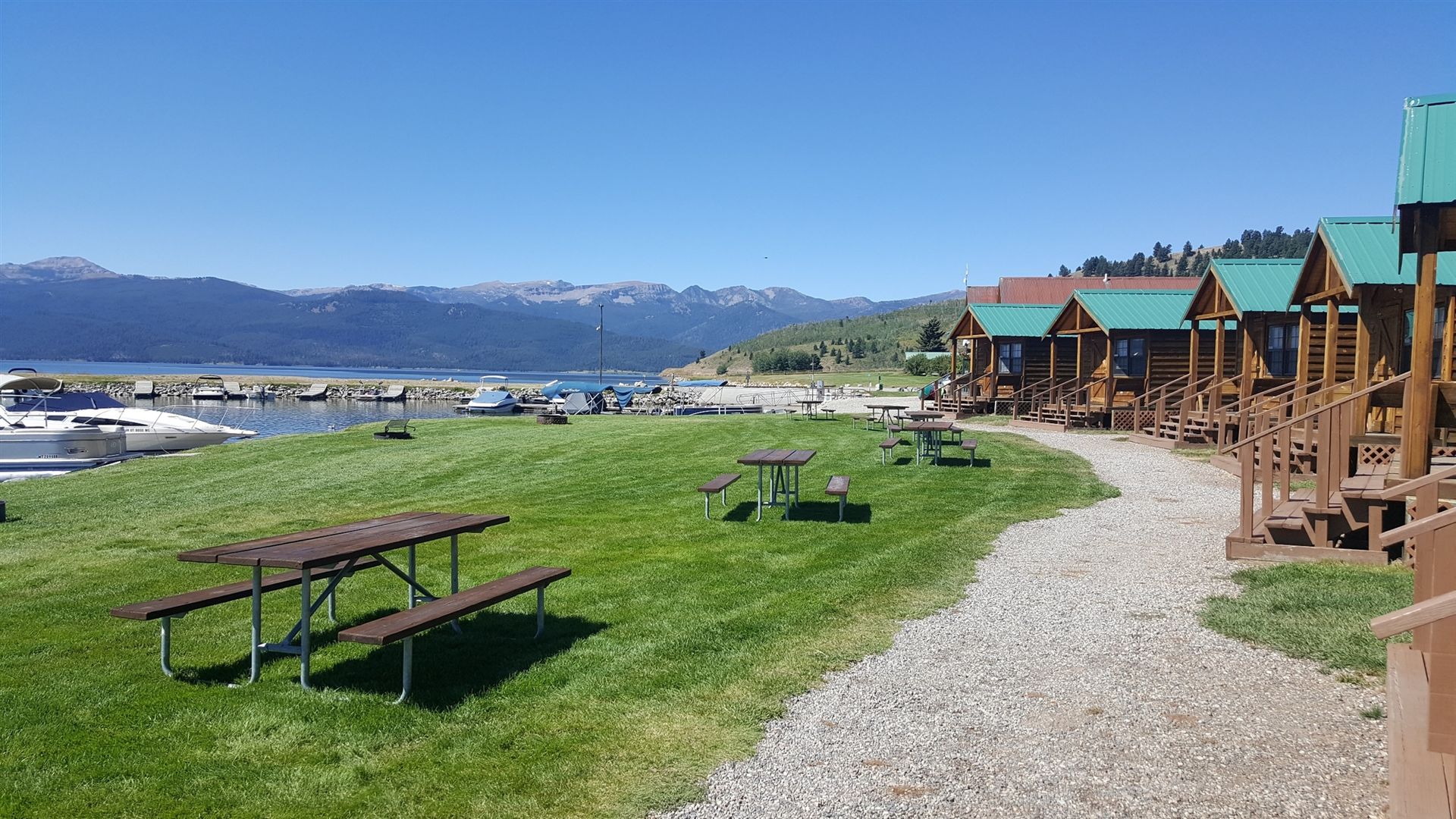 Lakeside picnic area with tables, gravel path, cabins, and mountains under a clear blue sky
