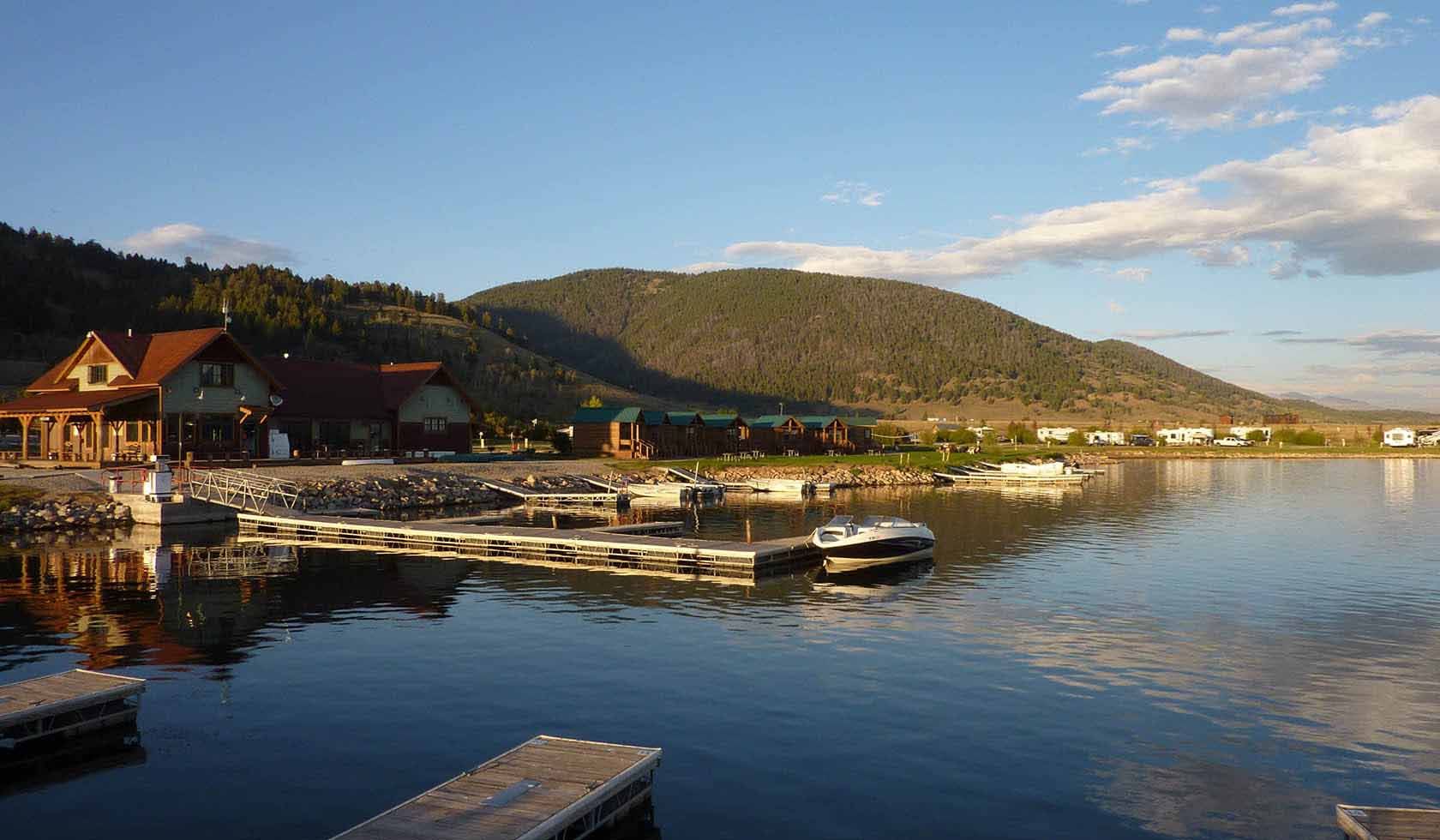 Calm lakeside marina at sunset with docked boats, cabins, and a forested hillside reflected in the water