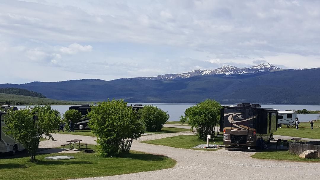 Lakeside park with trees, gravel path, picnic table, and snow-capped mountains under a clear sky