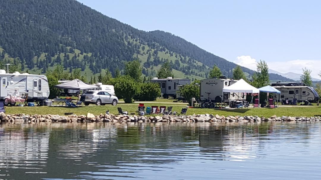 Riverside RV campground with trailers, tents, and a mountain backdrop beside calm water