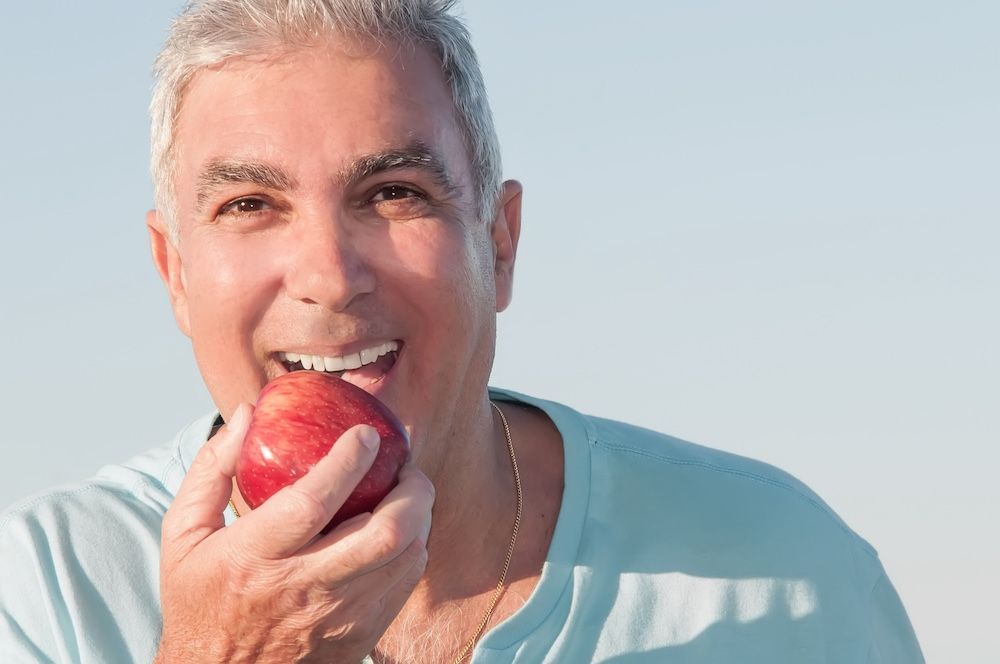 Man eating a red apple with an open mouth, blue shirt, bright background.