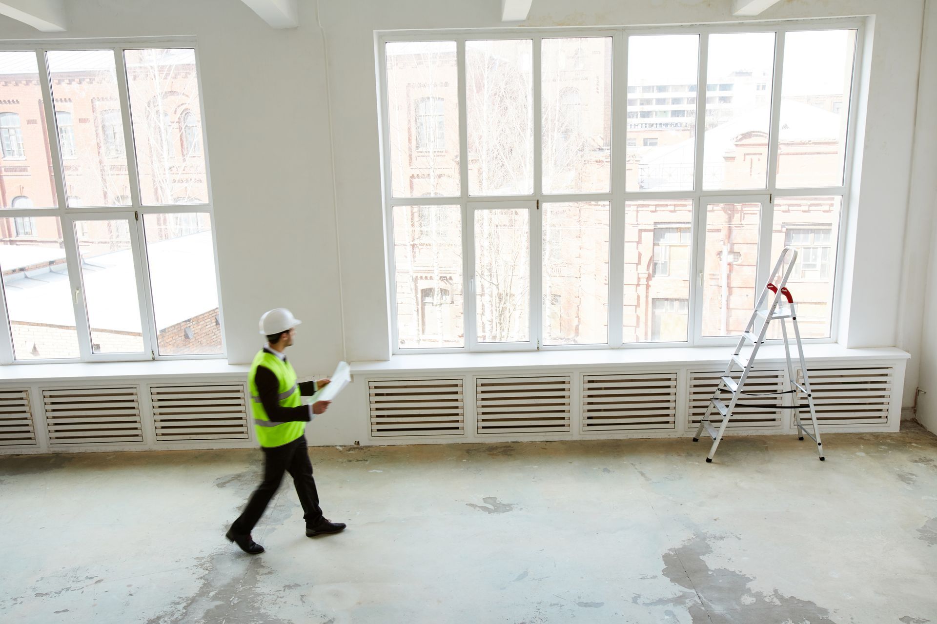 Worker in a high-visibility vest and hard hat walking through a bright room with large windows and a ladder.