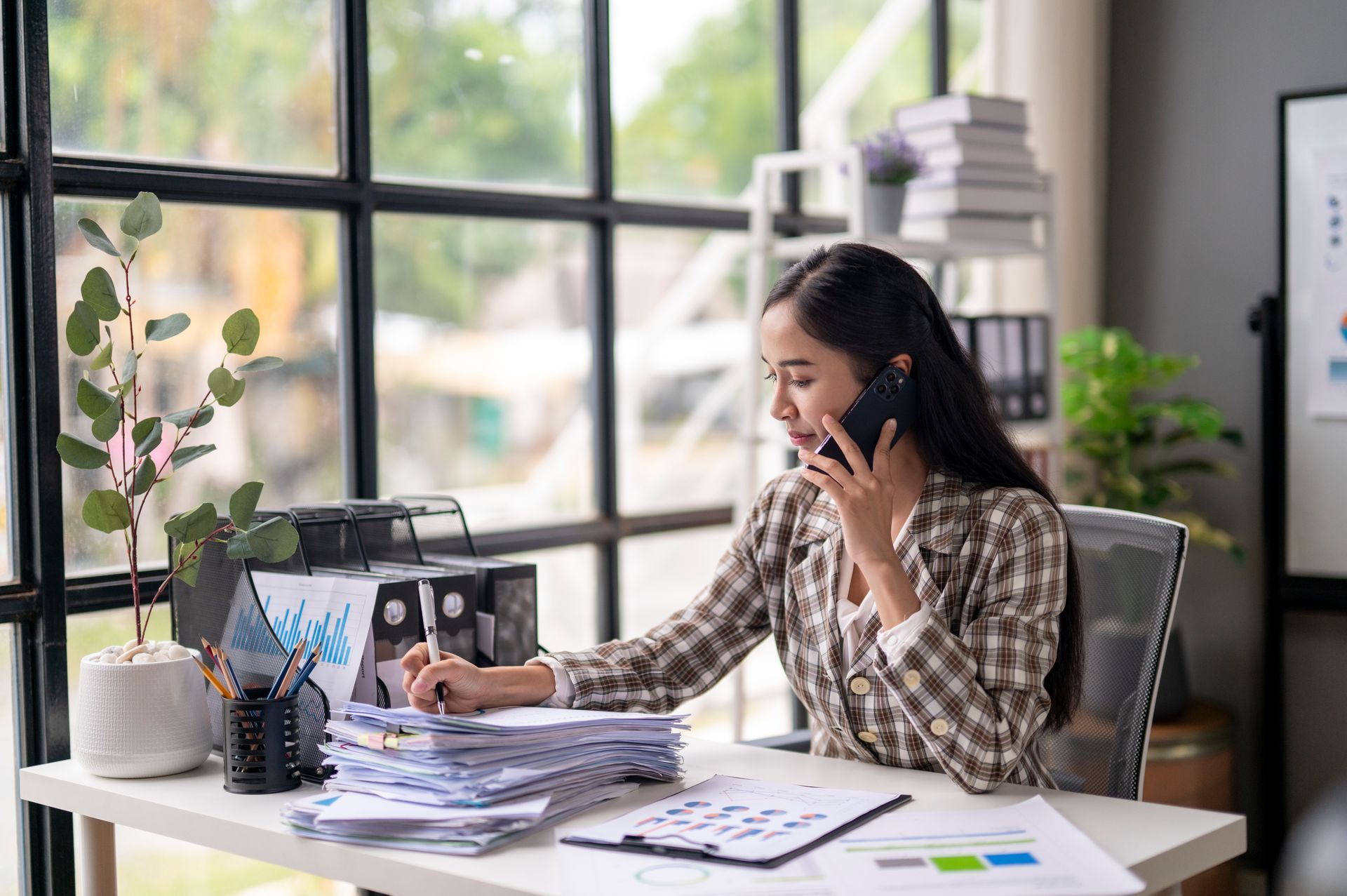 Woman working and talking on a phone at a desk in a bright office, with papers and a laptop.