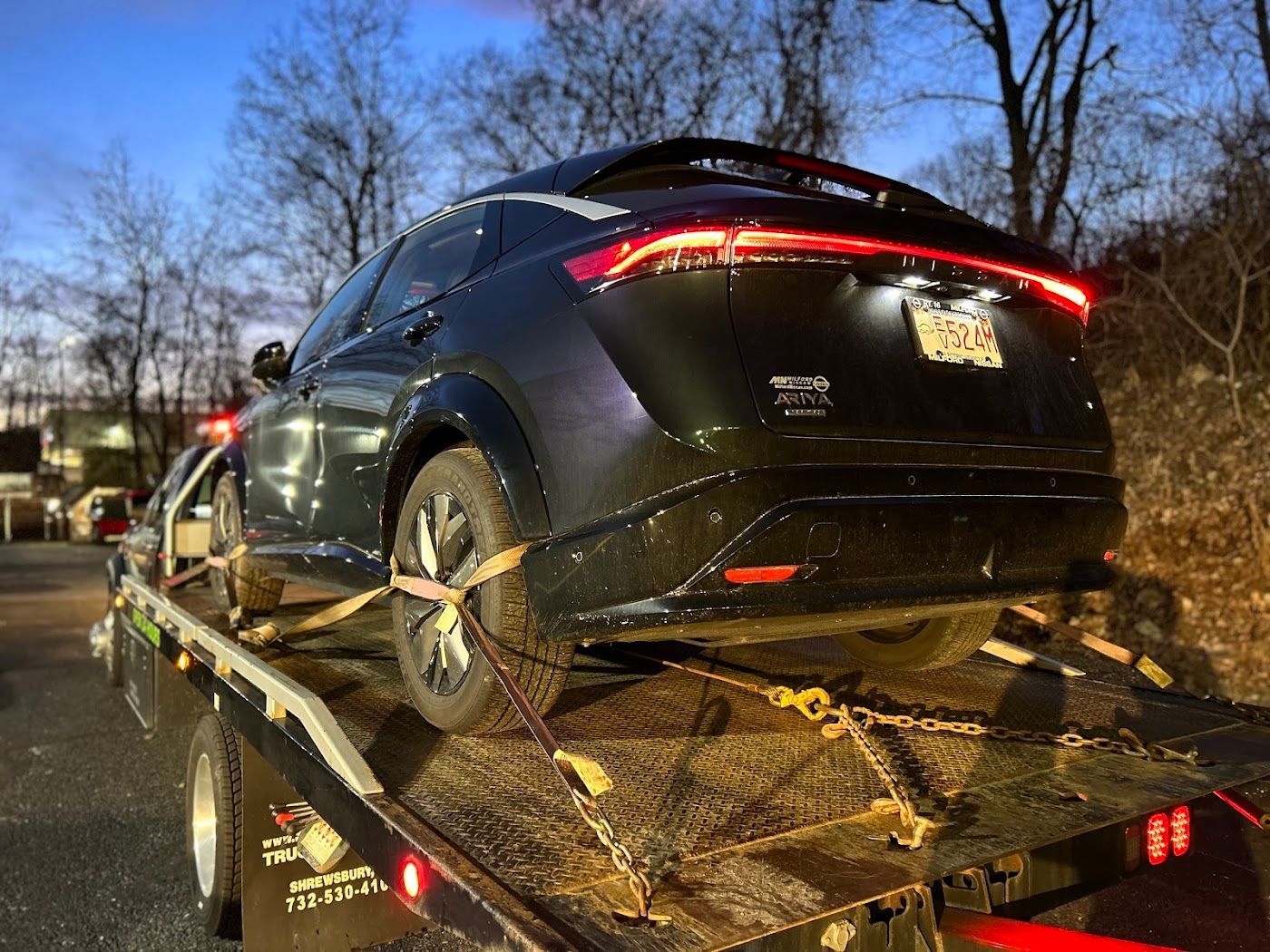 Dark car on a tow truck, rear view. Chains and straps secure the vehicle. Dusk setting with leafless trees.