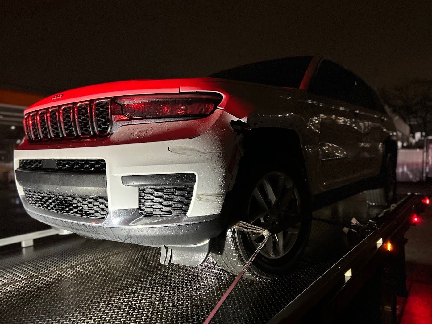 White SUV on a tow truck at night, lights illuminating the front grill and damaged bumper.