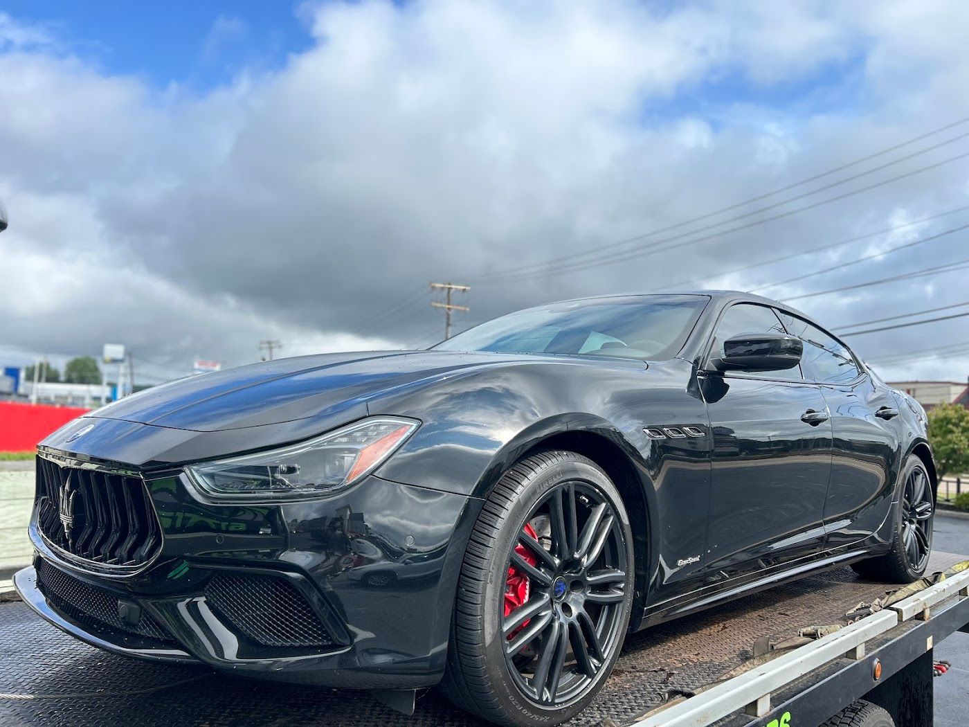 Black Maserati Ghibli on a tow truck, red brake calipers, cloudy sky.