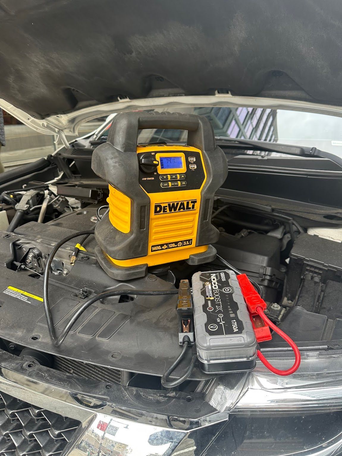 Yellow and black DeWalt car jump starter on a car hood, jumper cables attached.