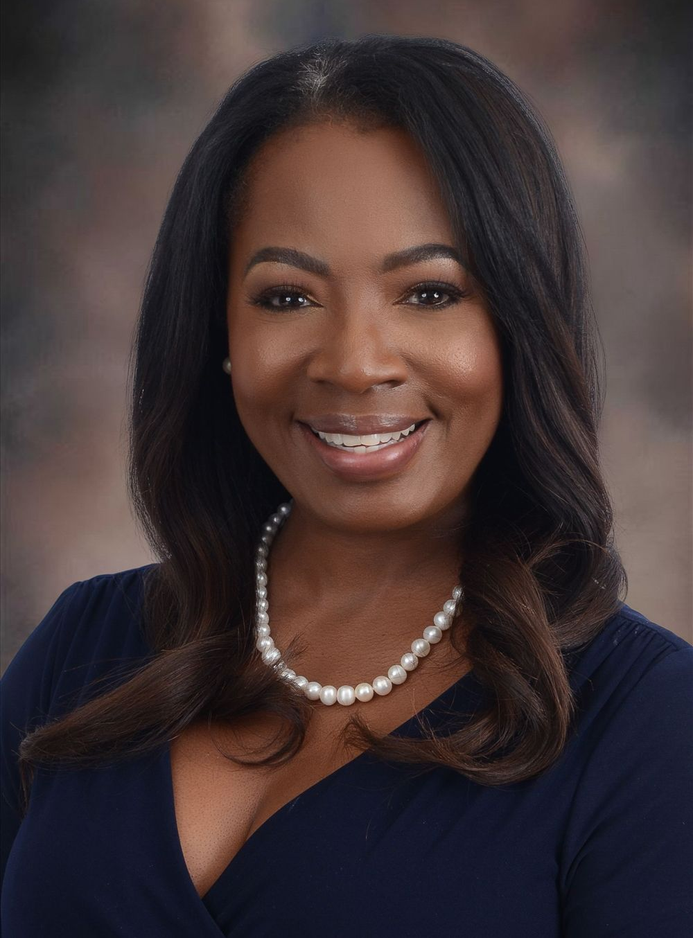 Woman with long dark hair smiles, wearing pearl necklace and blue top.