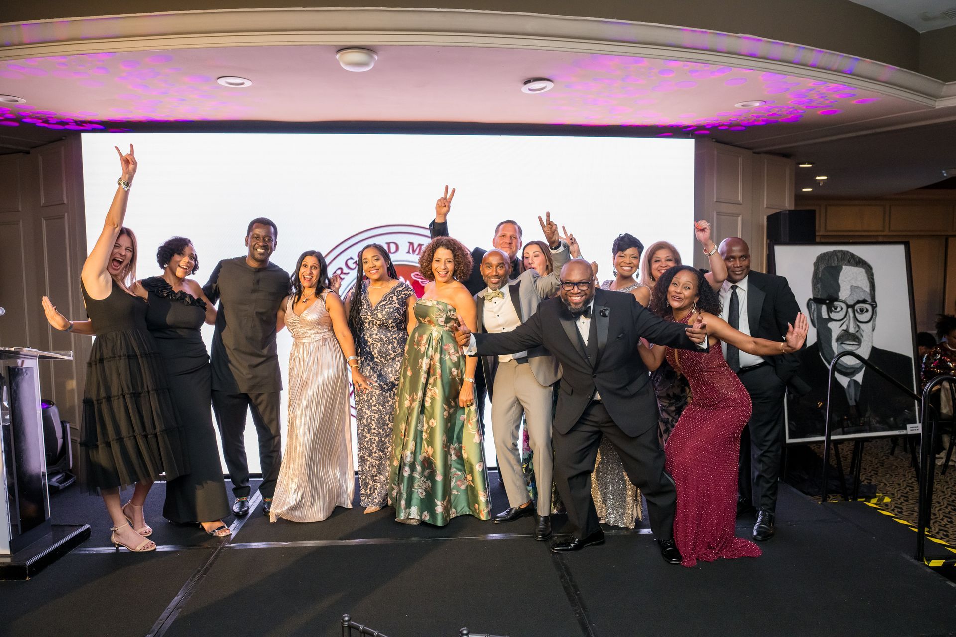 Group of people celebrating on stage in front of a screen with a logo, some holding up hands.