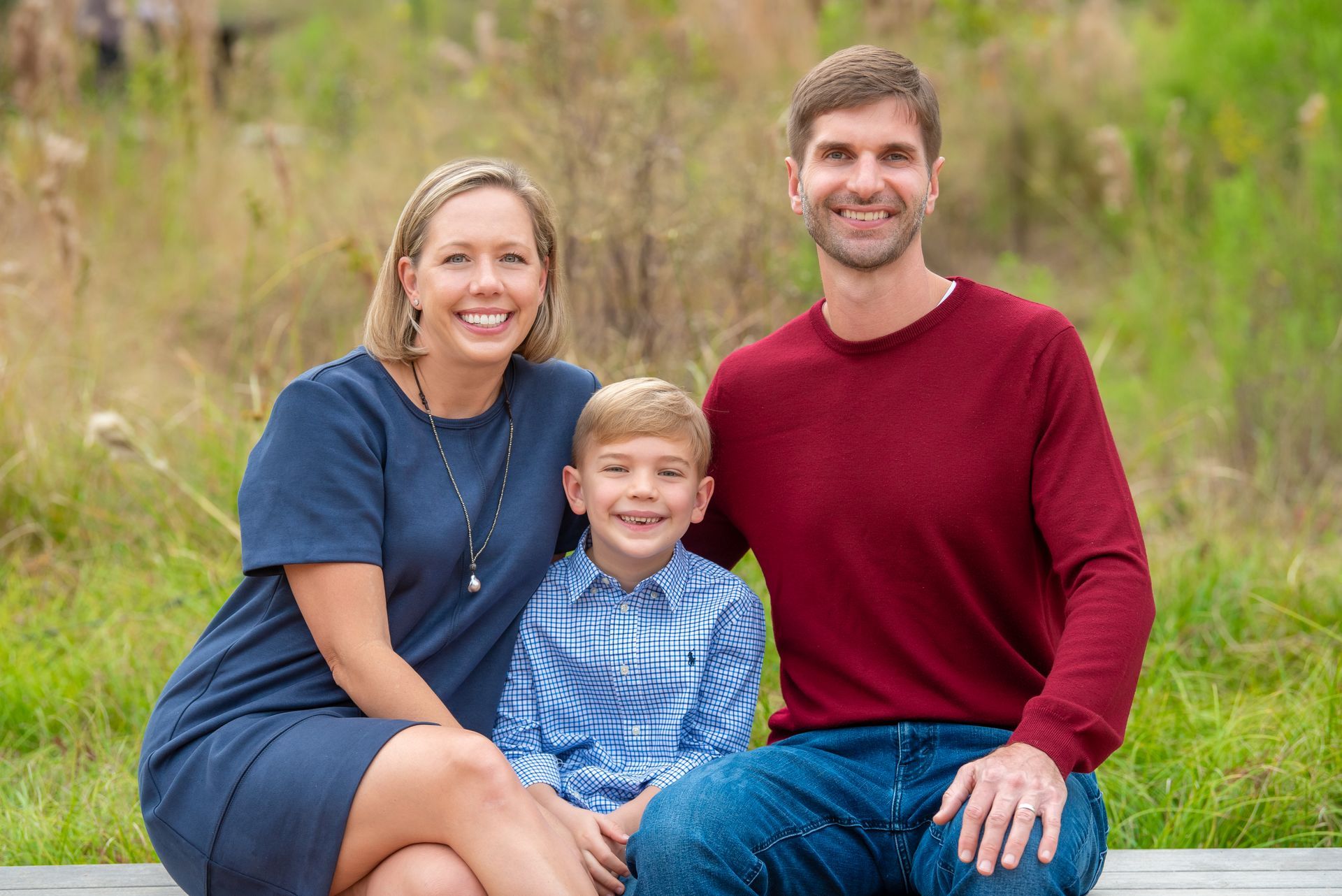 Family of three smiling, posing outdoors on a bench. Woman in blue dress, man in red shirt, child in patterned shirt.