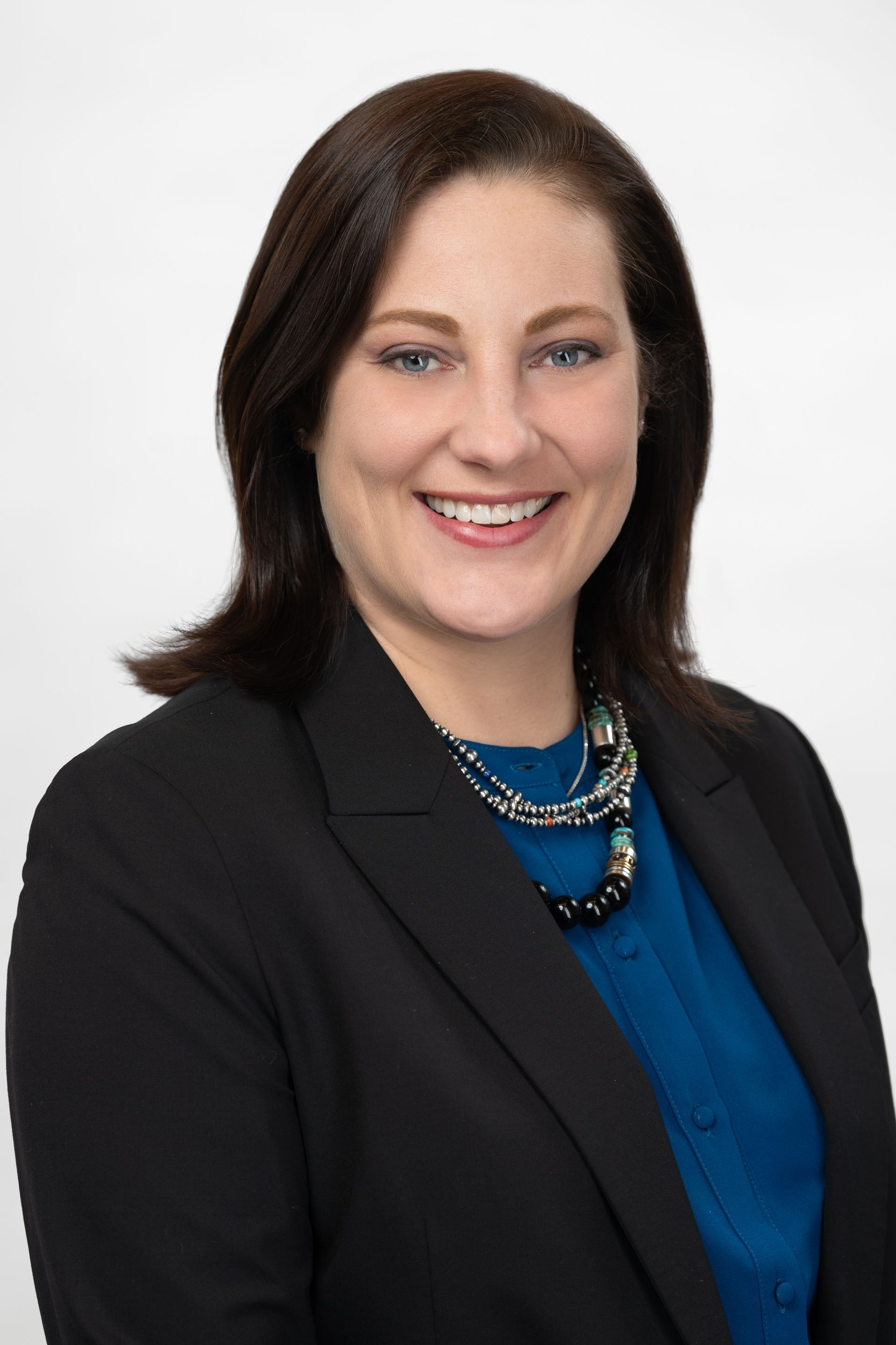 Woman with dark hair, smiling, wearing a blue blouse, black blazer, and beaded necklace. White background.