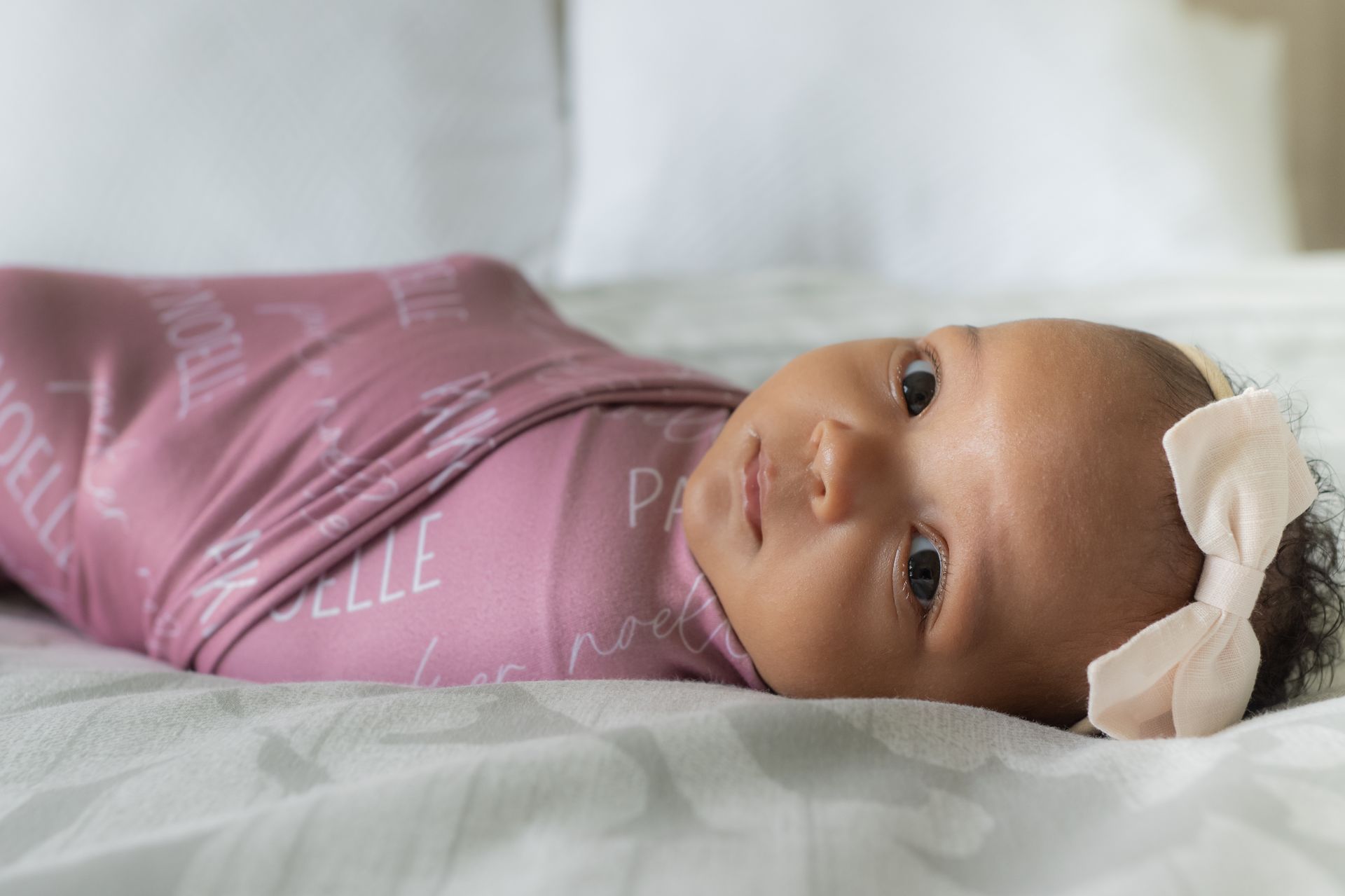 Baby swaddled in pink blanket with white text, wearing a bow, lying on a bed.