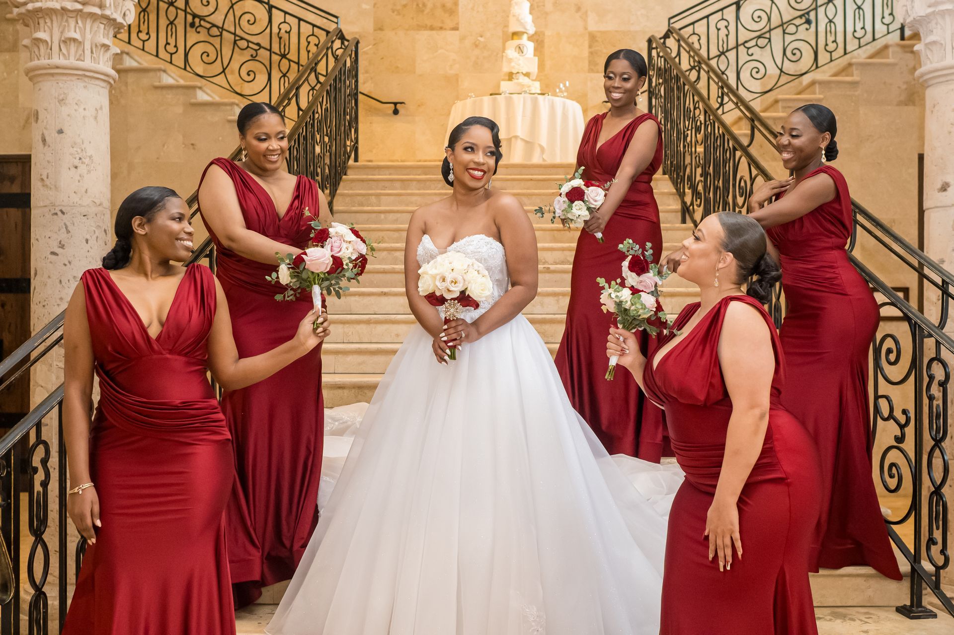 Bride with bridesmaids on stairs, wearing red gowns. Bride in white dress, holding bouquet, smiling.
