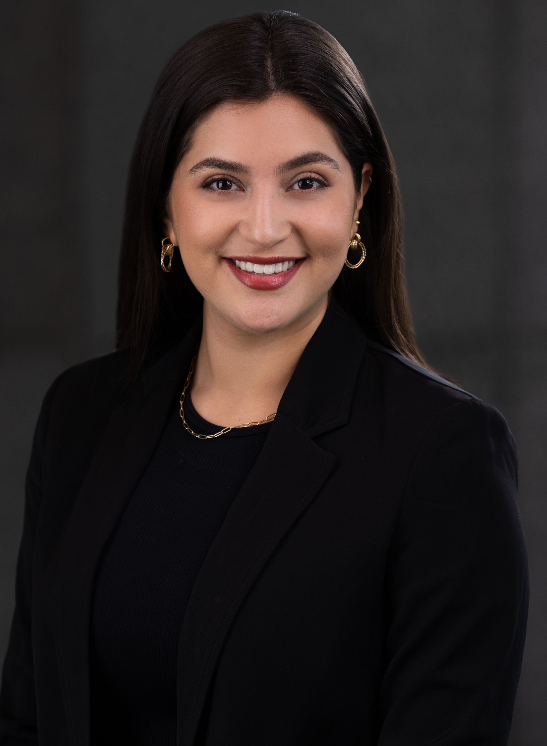 Woman in a black blazer and top smiling at the camera; dark hair, gold jewelry.