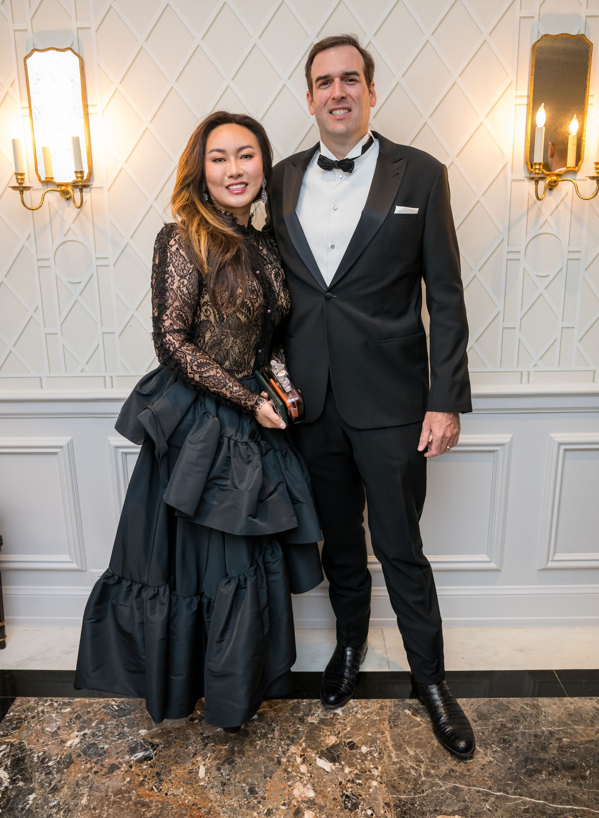 A formally dressed couple poses indoors. The woman wears a black gown, the man a tuxedo.
