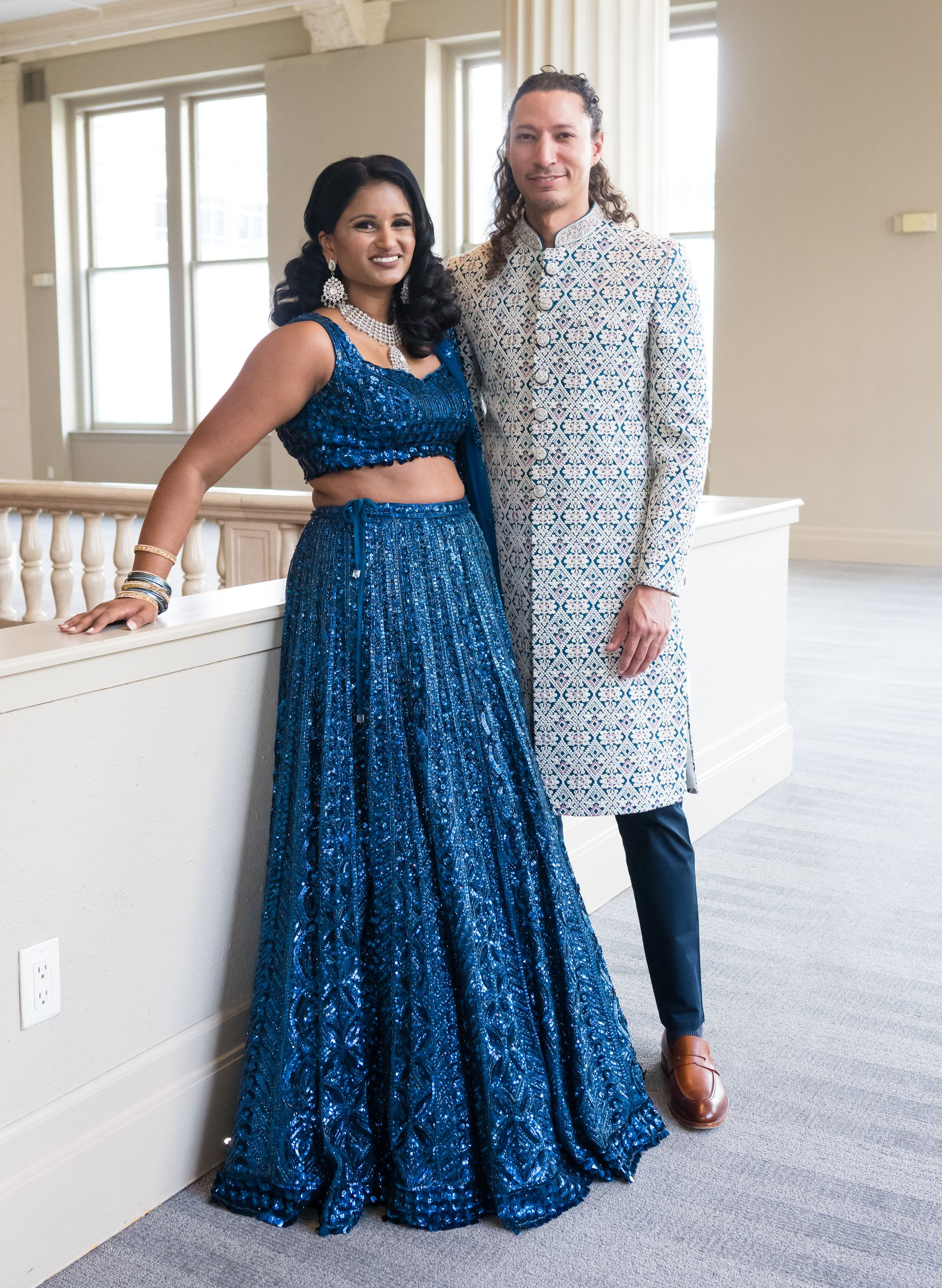 Couple in formal wear poses by a railing; woman in blue sequined outfit, man in patterned jacket.