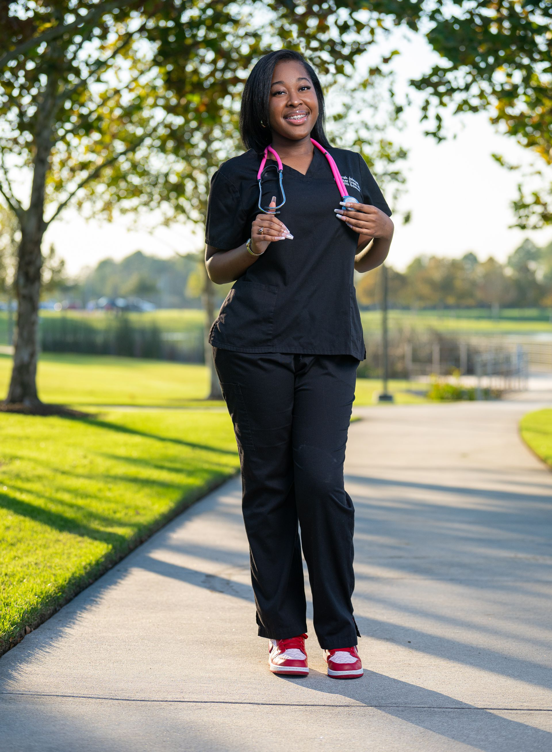 Person in black scrubs, pink stethoscope, walking on a path in a park, smiling.