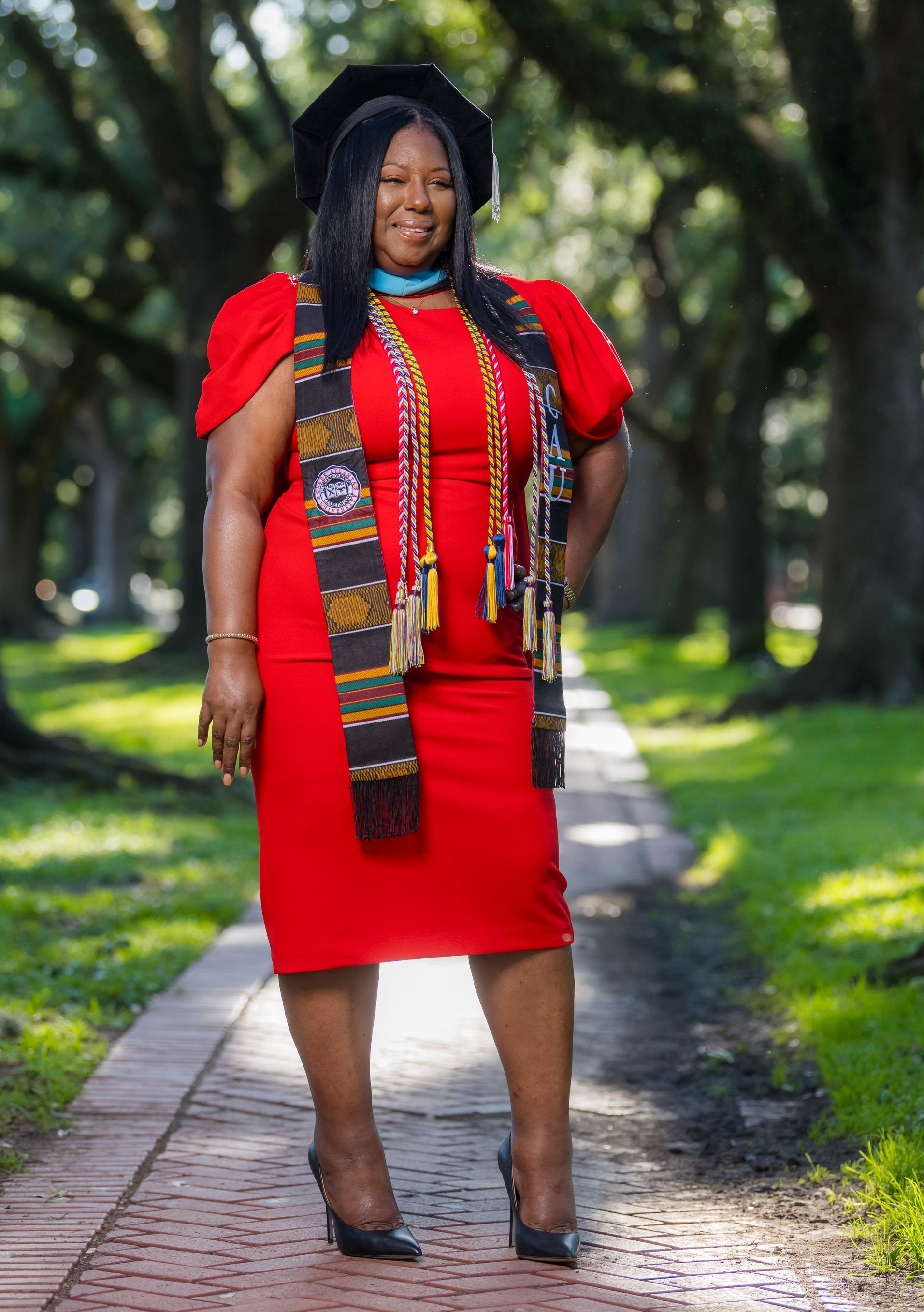 Woman in red dress and graduation attire, smiling outdoors on a brick path, trees in the background.