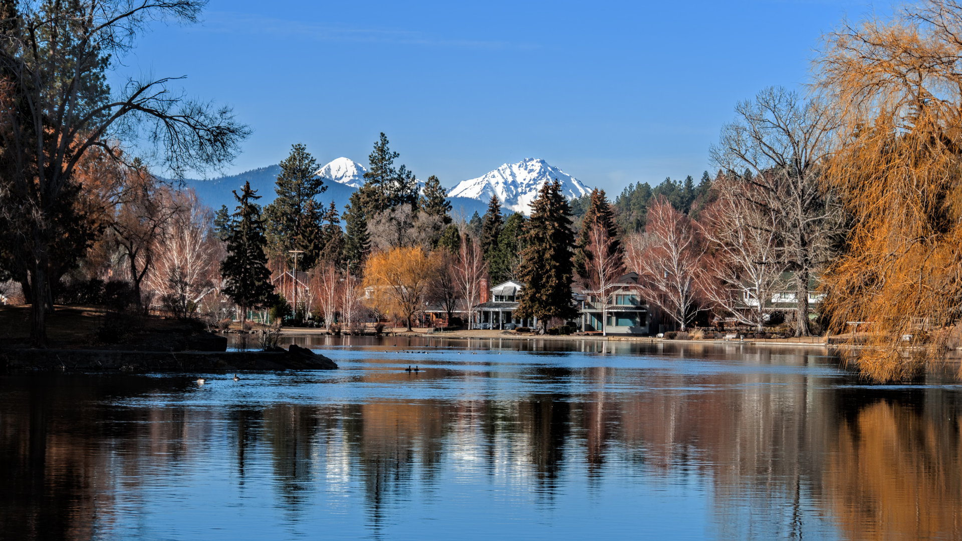 Snow-capped mountain reflected in a calm lake, with autumn trees and clear blue sky.